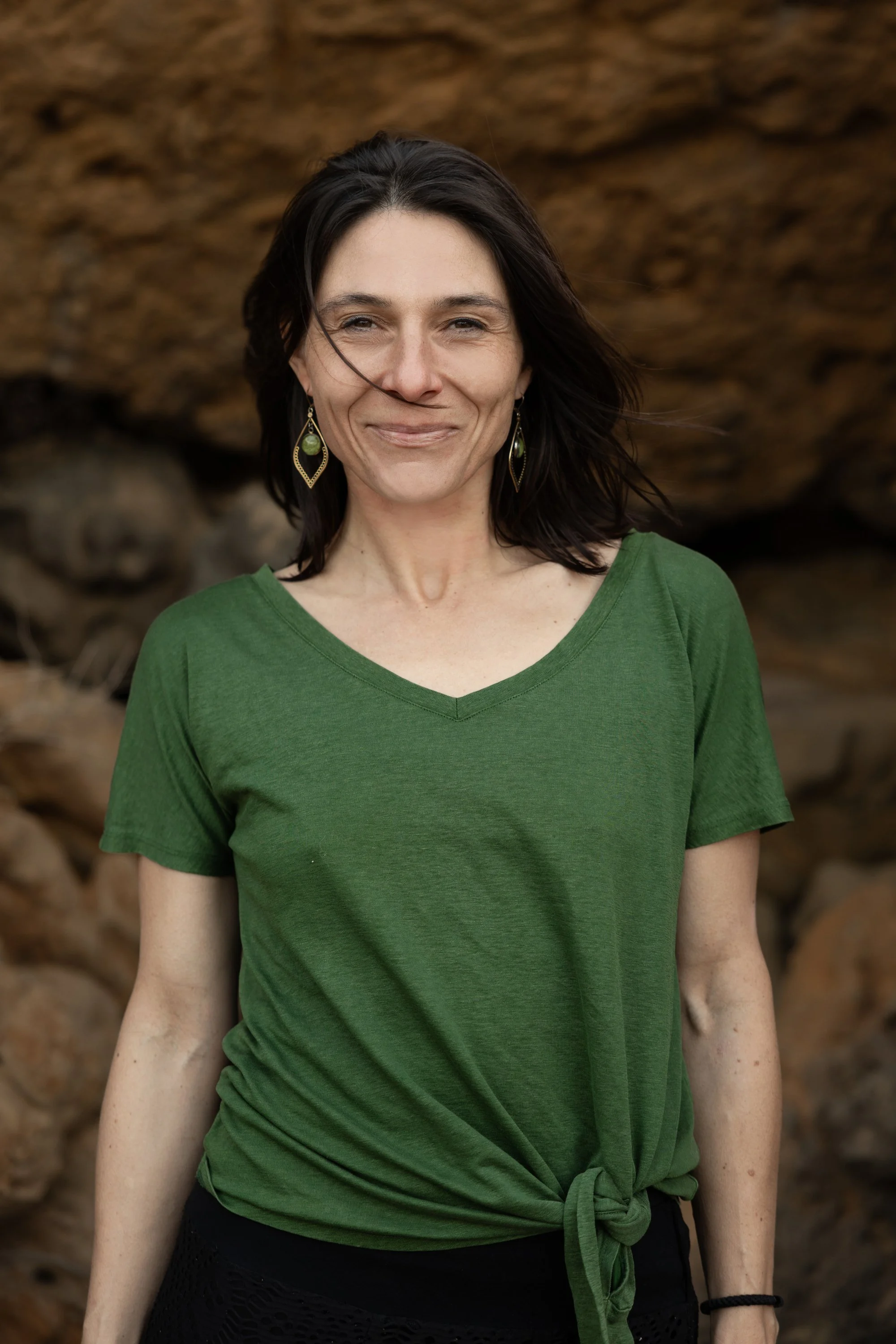 A woman with dark hair, wearing a green V-neck T-shirt tied at the waist, standing outdoors with a rocky background.
