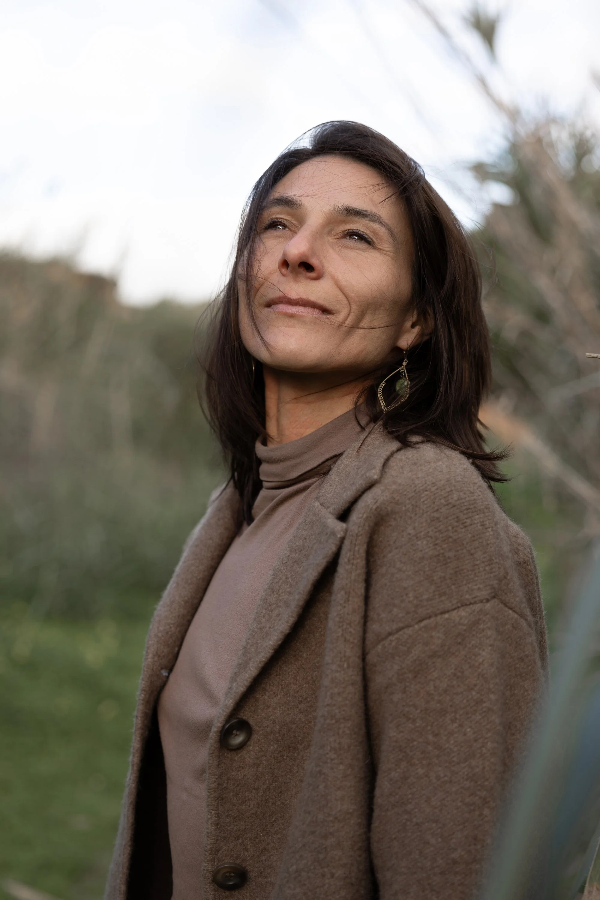 A woman with dark hair and earrings, wearing a brown coat and tan turtleneck, outdoors with a blurred natural background.