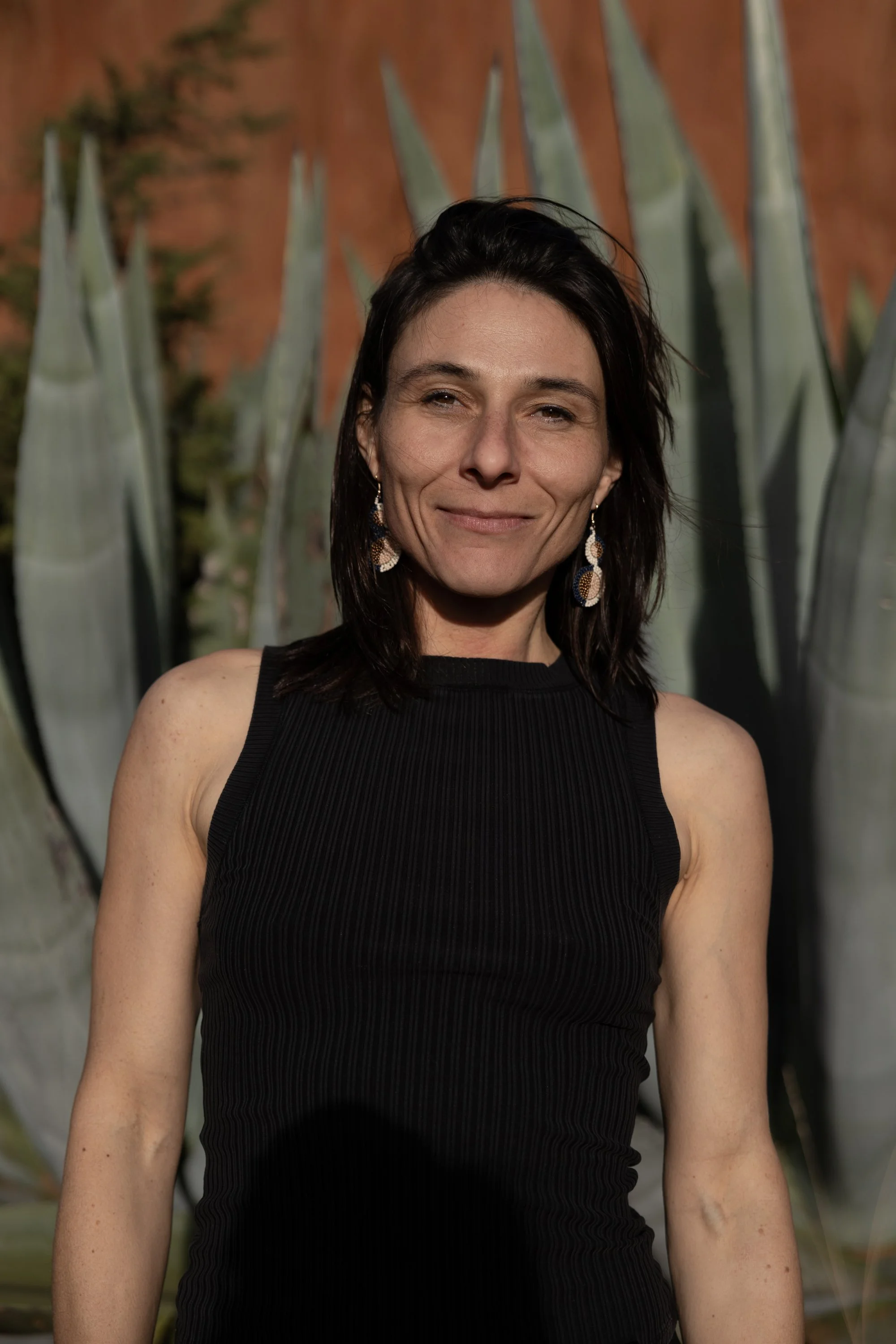 A woman with dark hair wearing a sleeveless black top, standing outdoors in front of a large agave plant with a reddish-brown wall behind it.