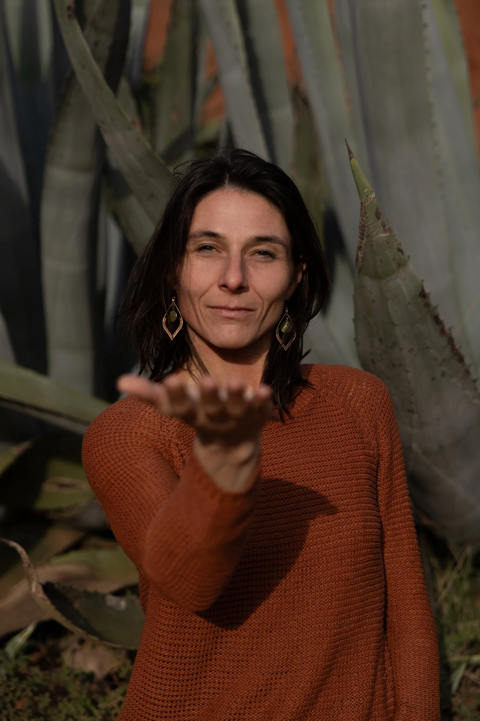 A woman with dark hair wearing a rust-colored sweater, standing outdoors in front of large agave plants, reaching out with her right hand towards the camera.