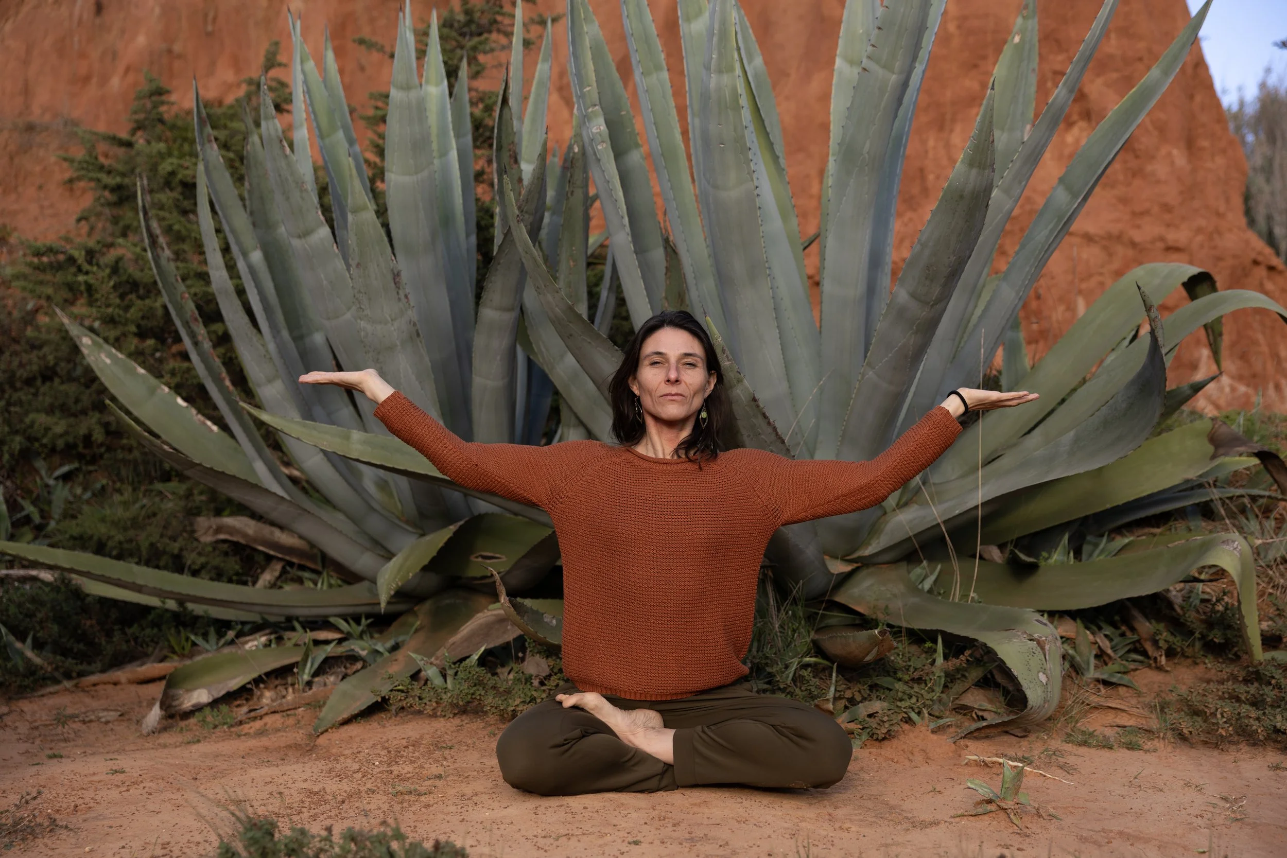A woman practicing yoga outdoors, seated cross-legged on the ground with a large agave plant behind her. She has her arms extended to the sides, palms facing upward, in a meditative pose.