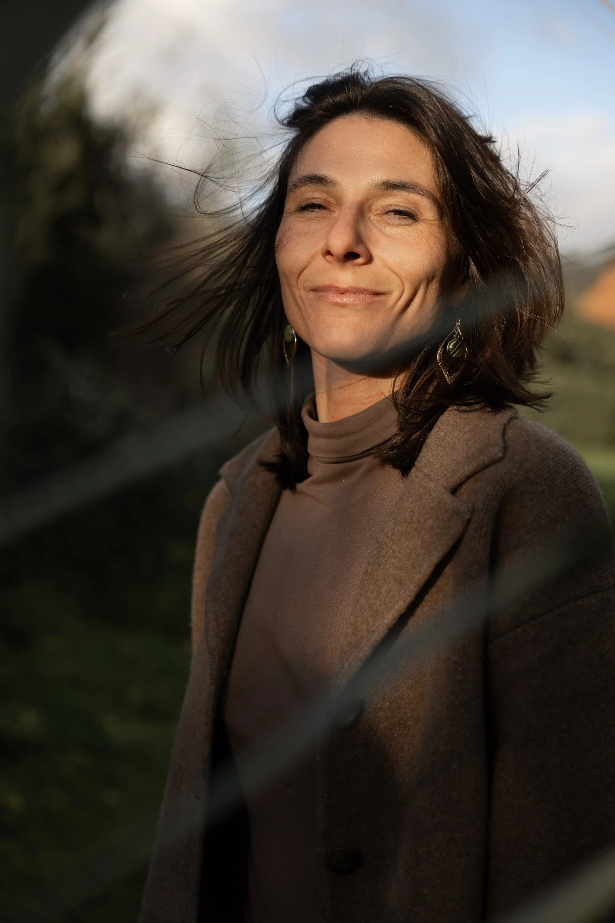 A woman with shoulder-length dark hair and earrings, wearing a brown blazer and turtleneck, standing outdoors with a confident expression, sunlight on her face, and a blurred background of greenery and sky.