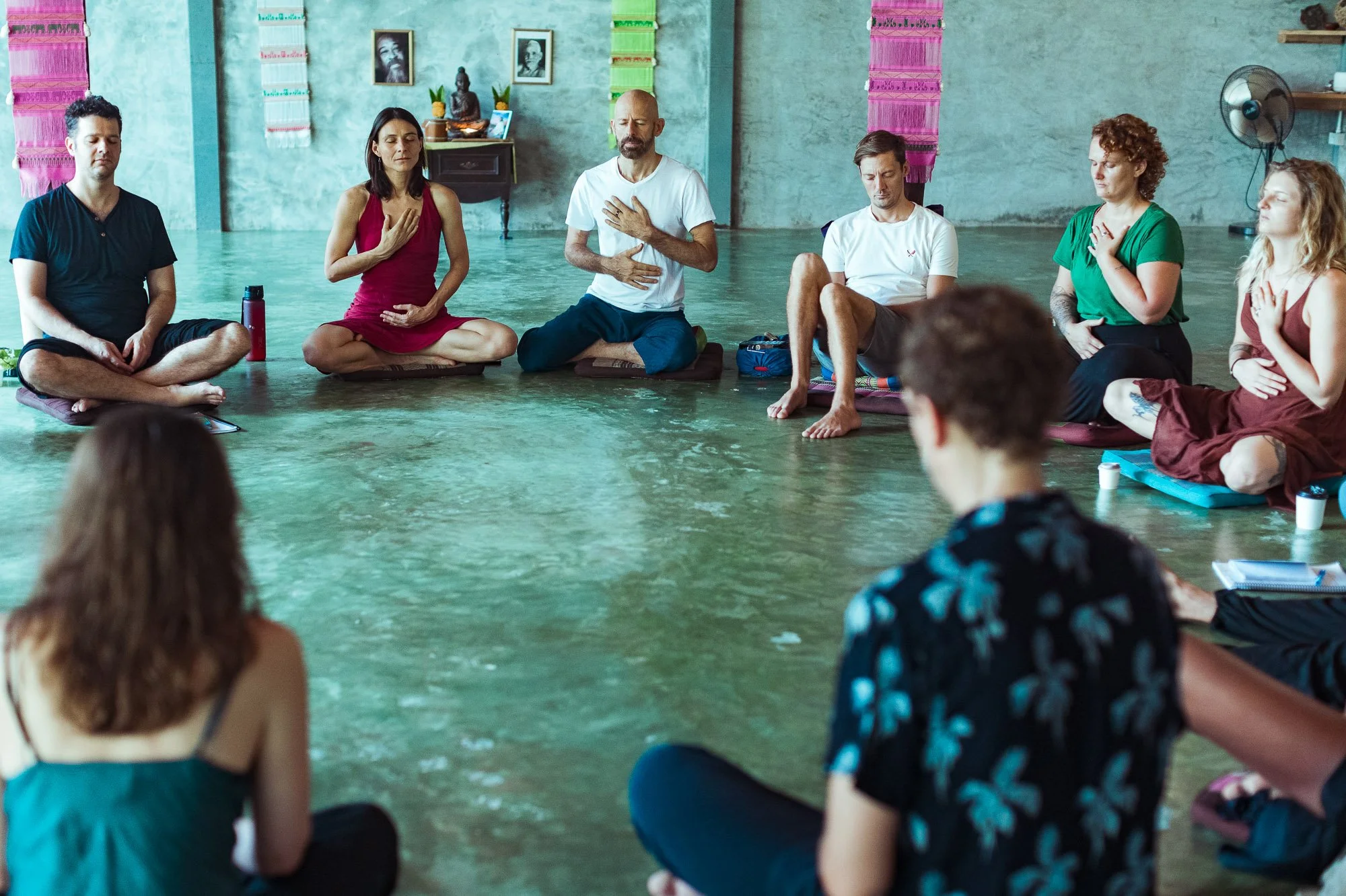 Group of people practicing meditation or yoga in a circle on the floor in a spacious room with teal walls, colorful wall hangings, and spiritual statues.