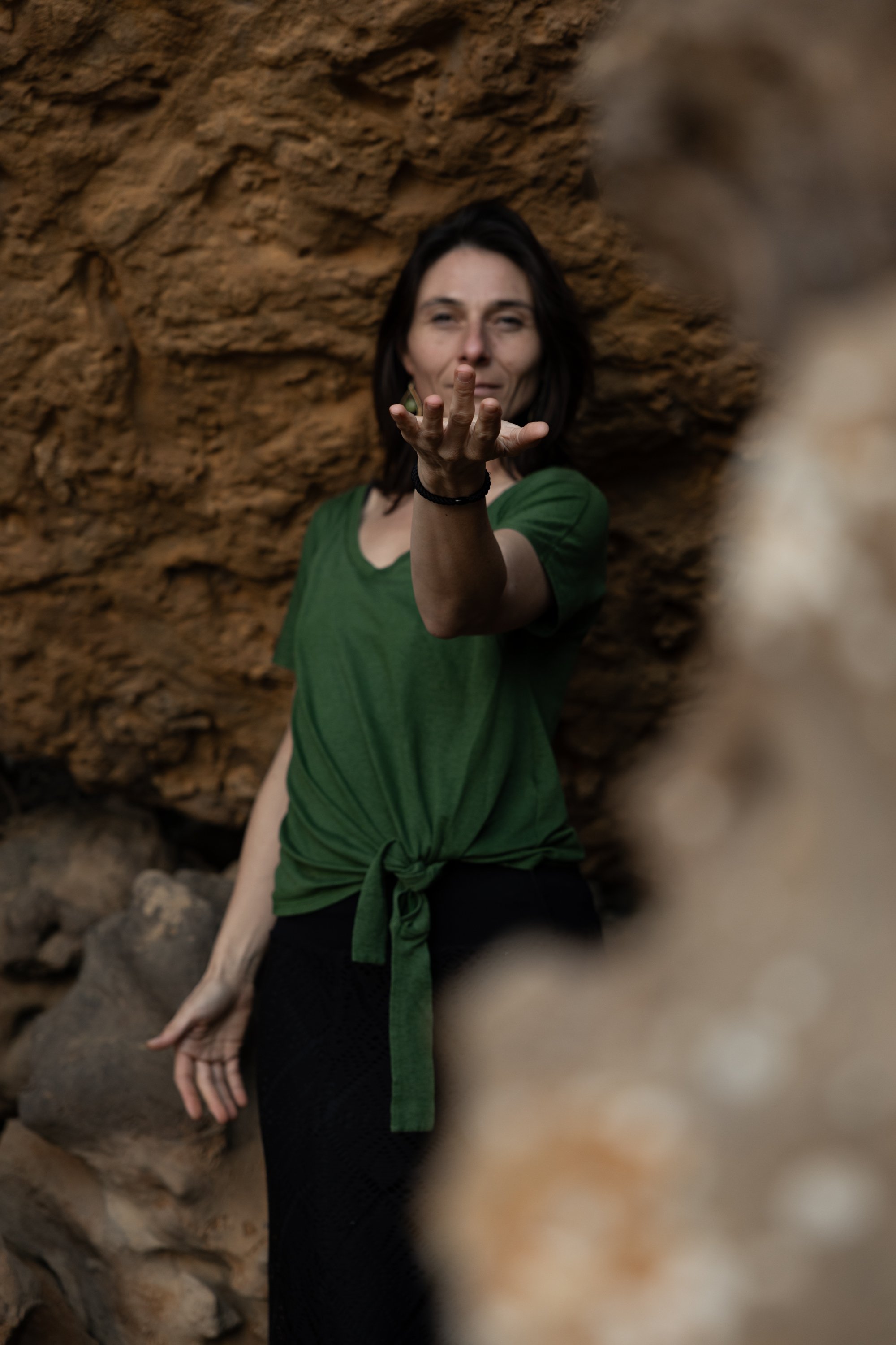 Woman wearing a green shirt extending her hand towards the camera in a rocky outdoor setting.