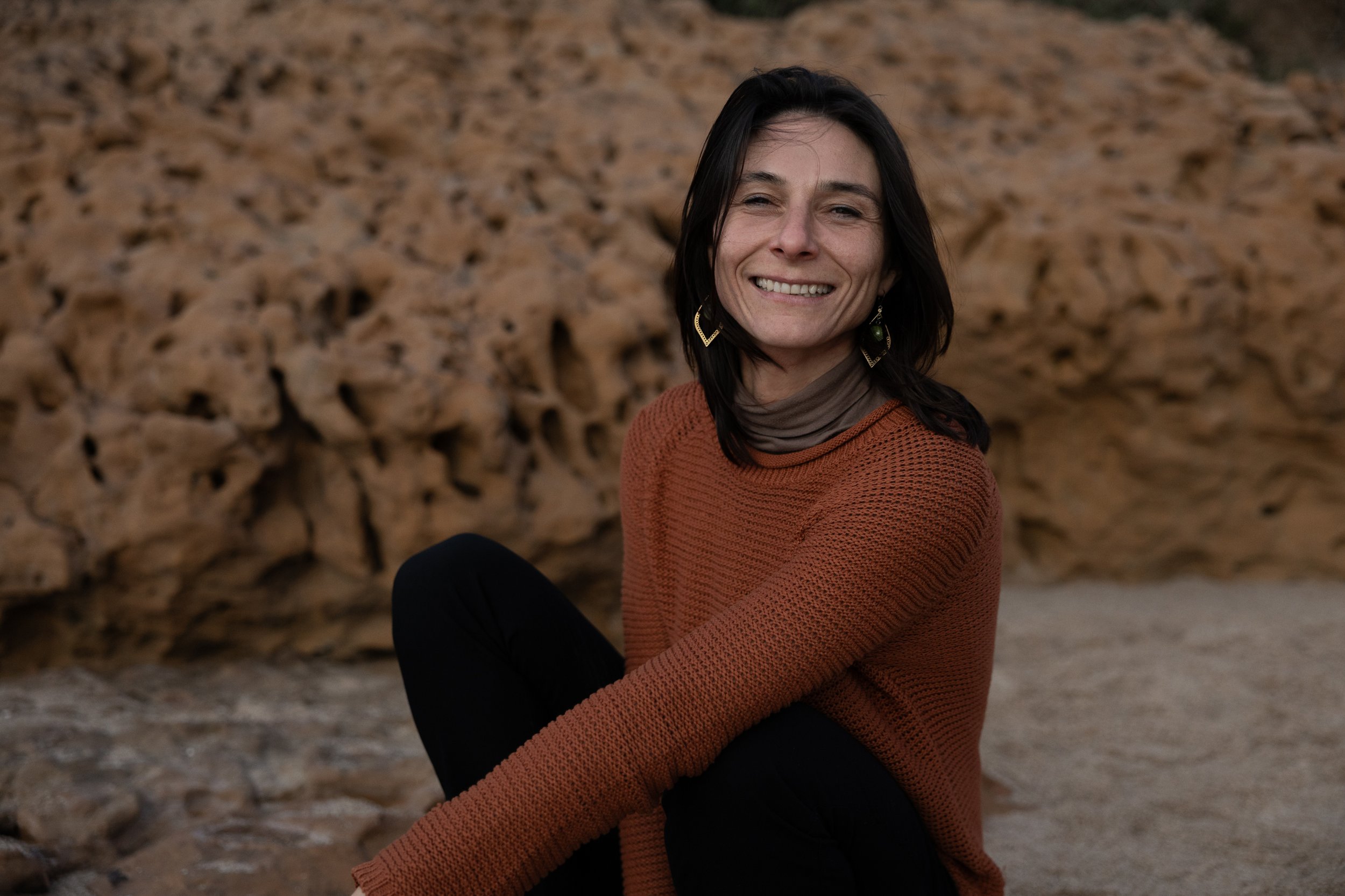 A woman with dark hair smiling, wearing a rust-colored sweater and black pants, sitting on the ground in front of rocky terrain.