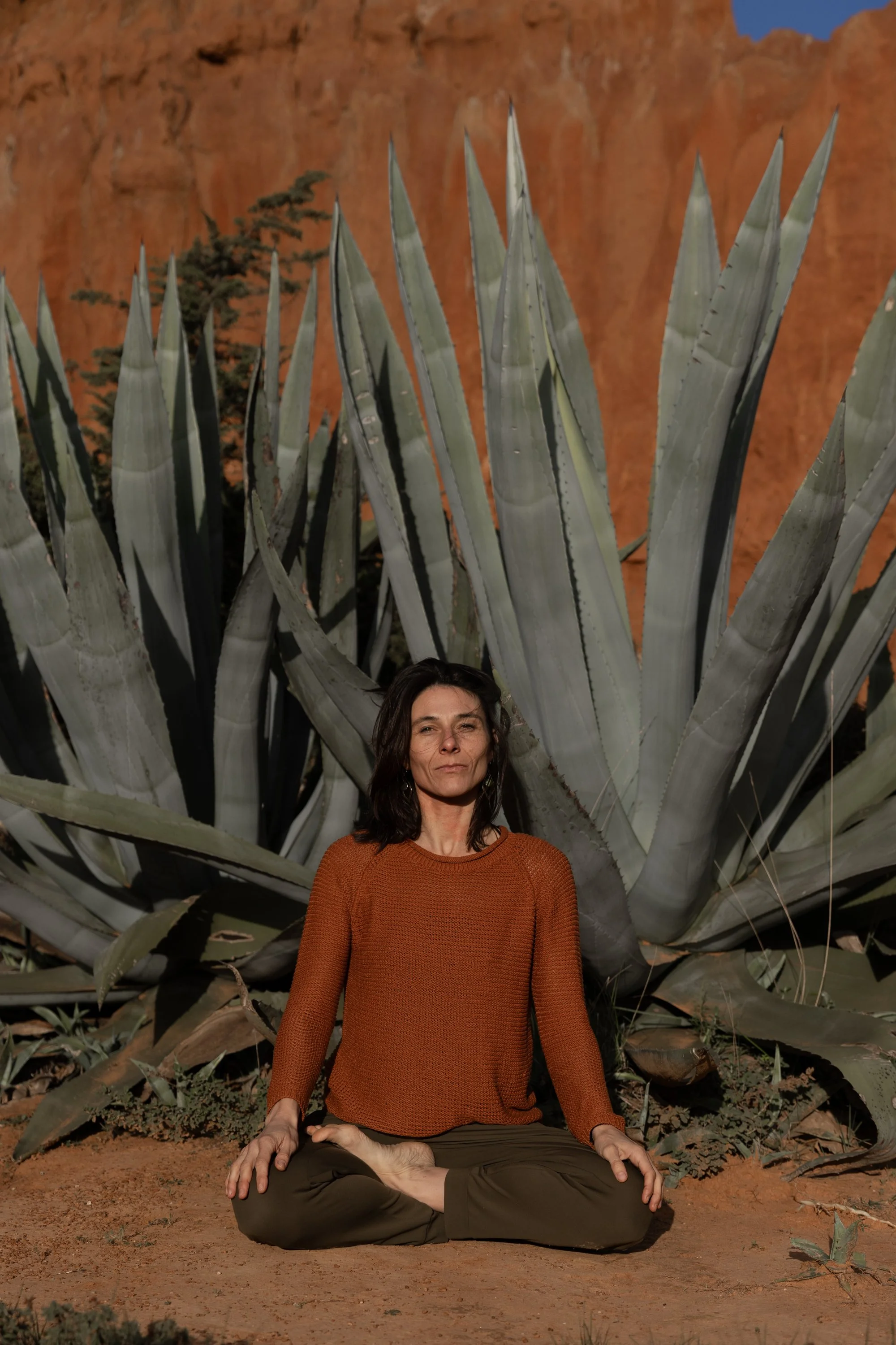 A woman in an orange sweater and brown pants is practicing yoga and meditation in a desert landscape, sitting cross-legged with her hands resting on her knees in front of an agave plant against a reddish-orange cliff background.