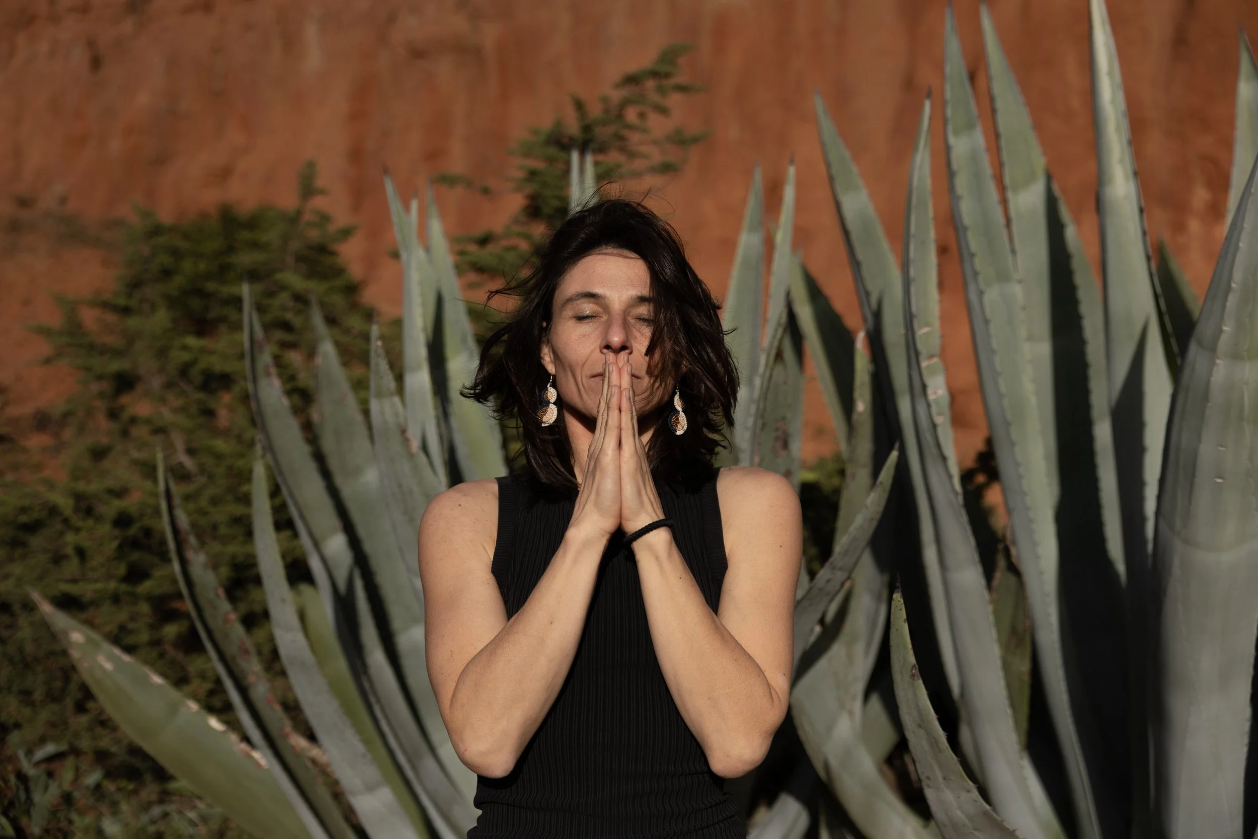 A woman with dark hair in a black sleeveless top stands with her eyes closed and hands pressed together in a prayer position, amidst large agave plants and a desert landscape with a reddish hill in the background.