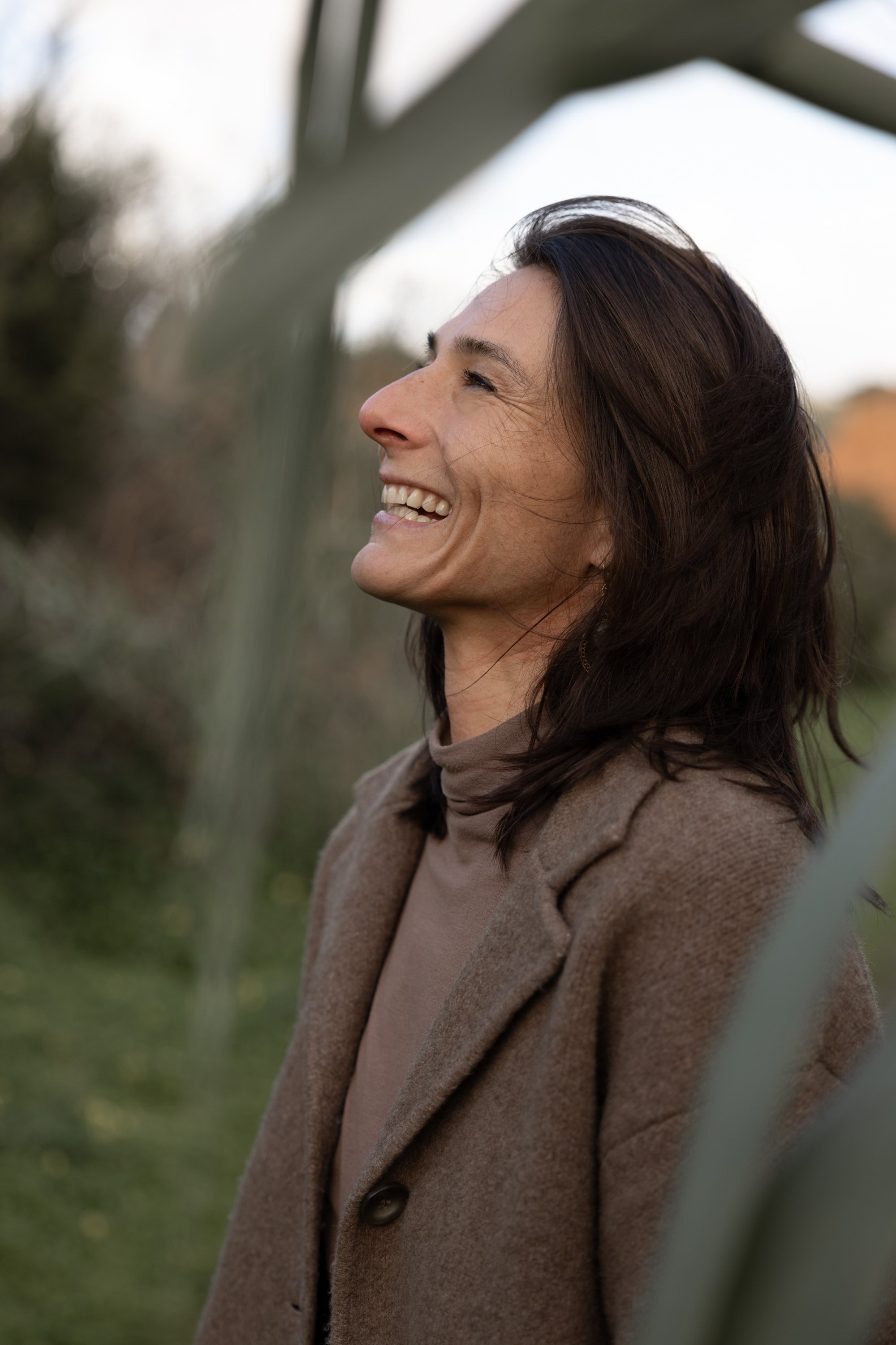 A woman with shoulder-length dark hair smiling outdoors during daytime, wearing a beige coat and top, with blurred trees and grass in the background.