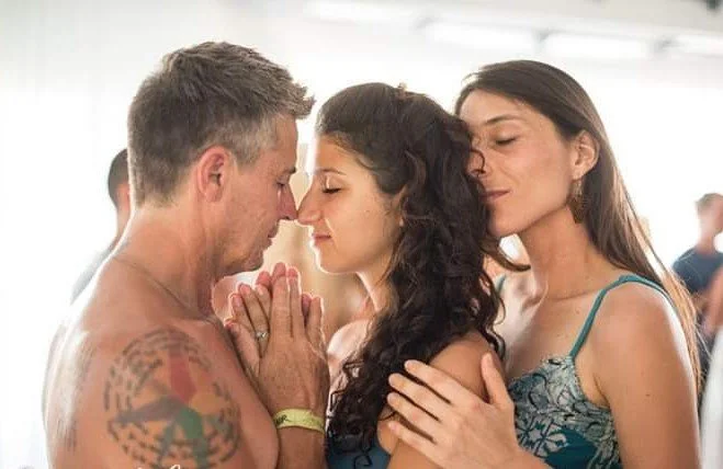 Three people in a close, intimate embrace with their foreheads touching, eyes closed, in a bright indoor setting.