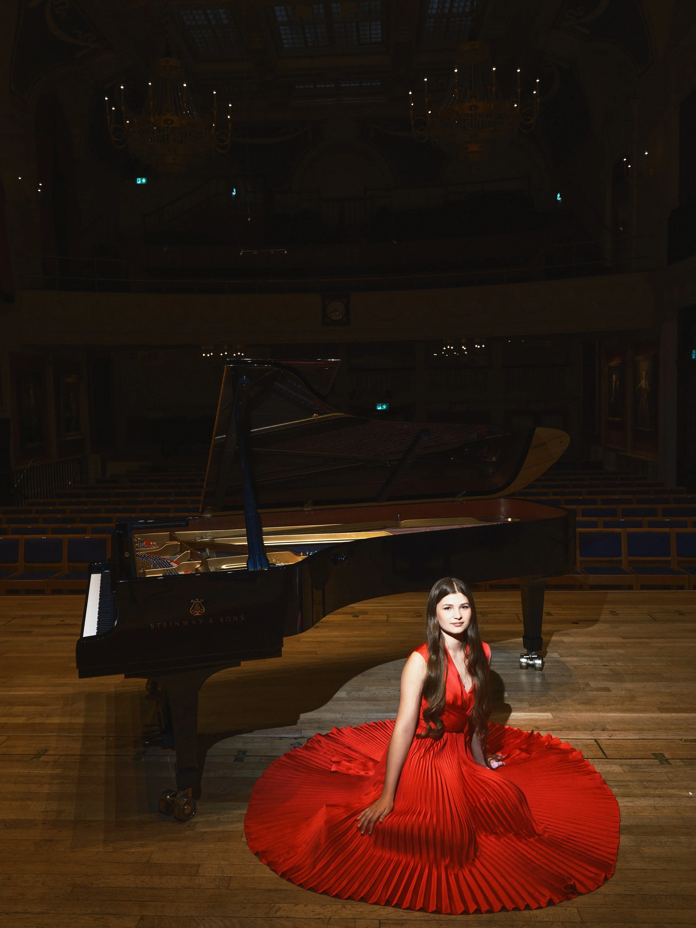 A young woman in a long red pleated dress sitting on a wooden stage in front of a grand piano in an empty concert hall.