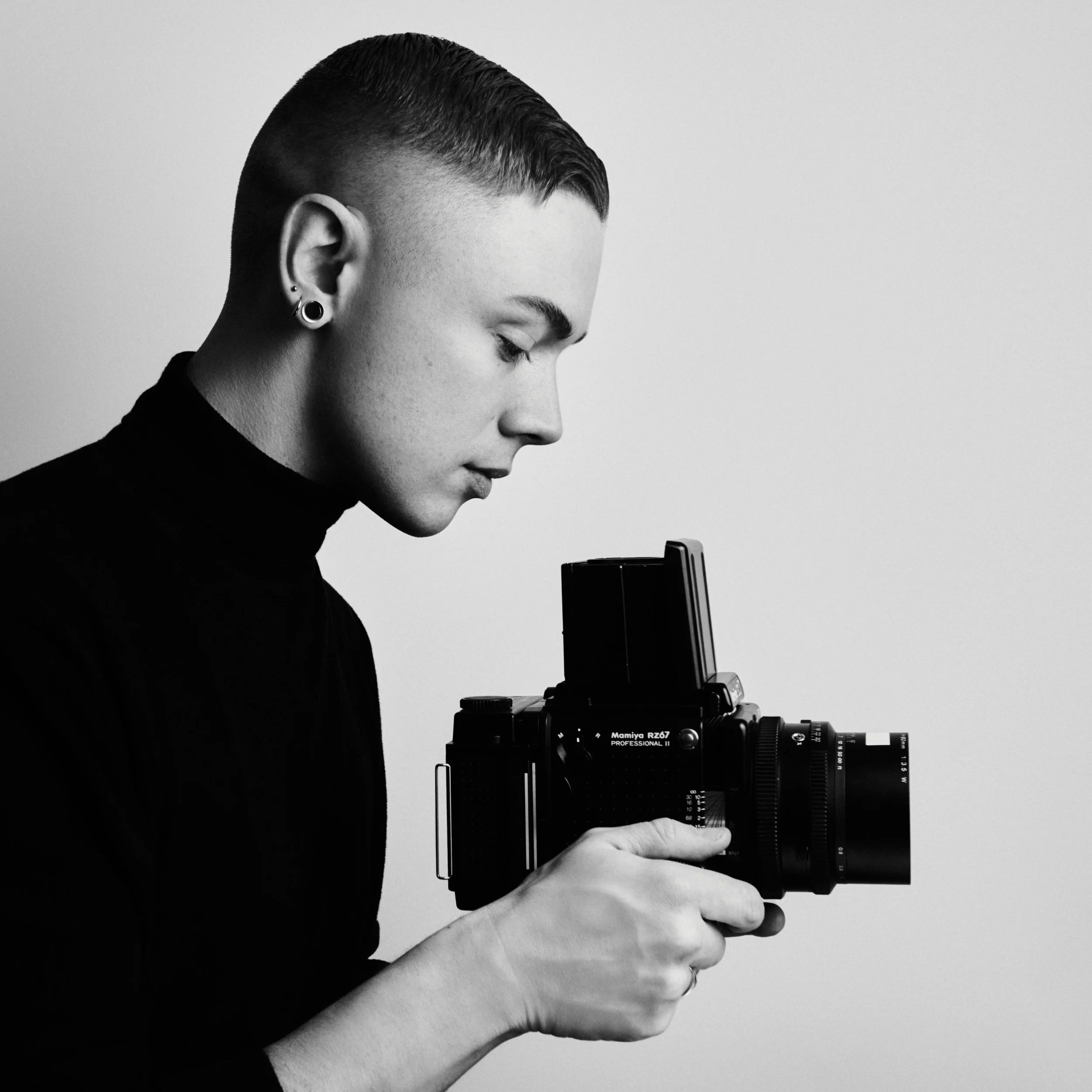 Black and white photo of a man with short hair, wearing earrings and a turtleneck, holding a camera and looking down.