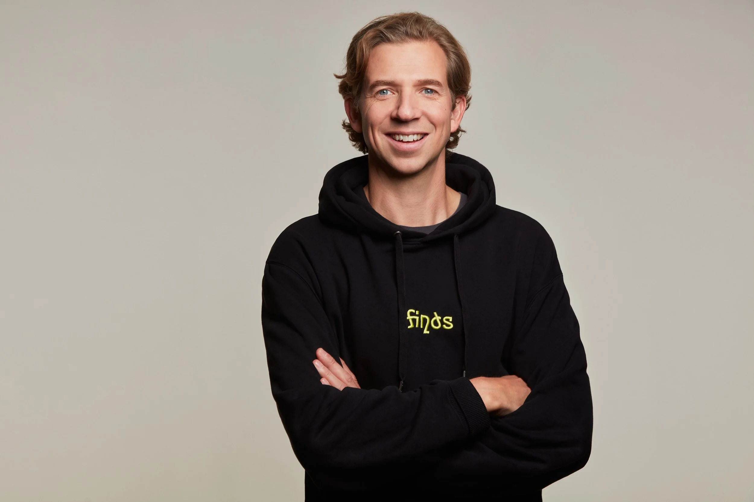 A smiling young man with blond hair wearing a black hoodie with yellow text that says 'finds', posing with arms crossed against a plain light gray background.
