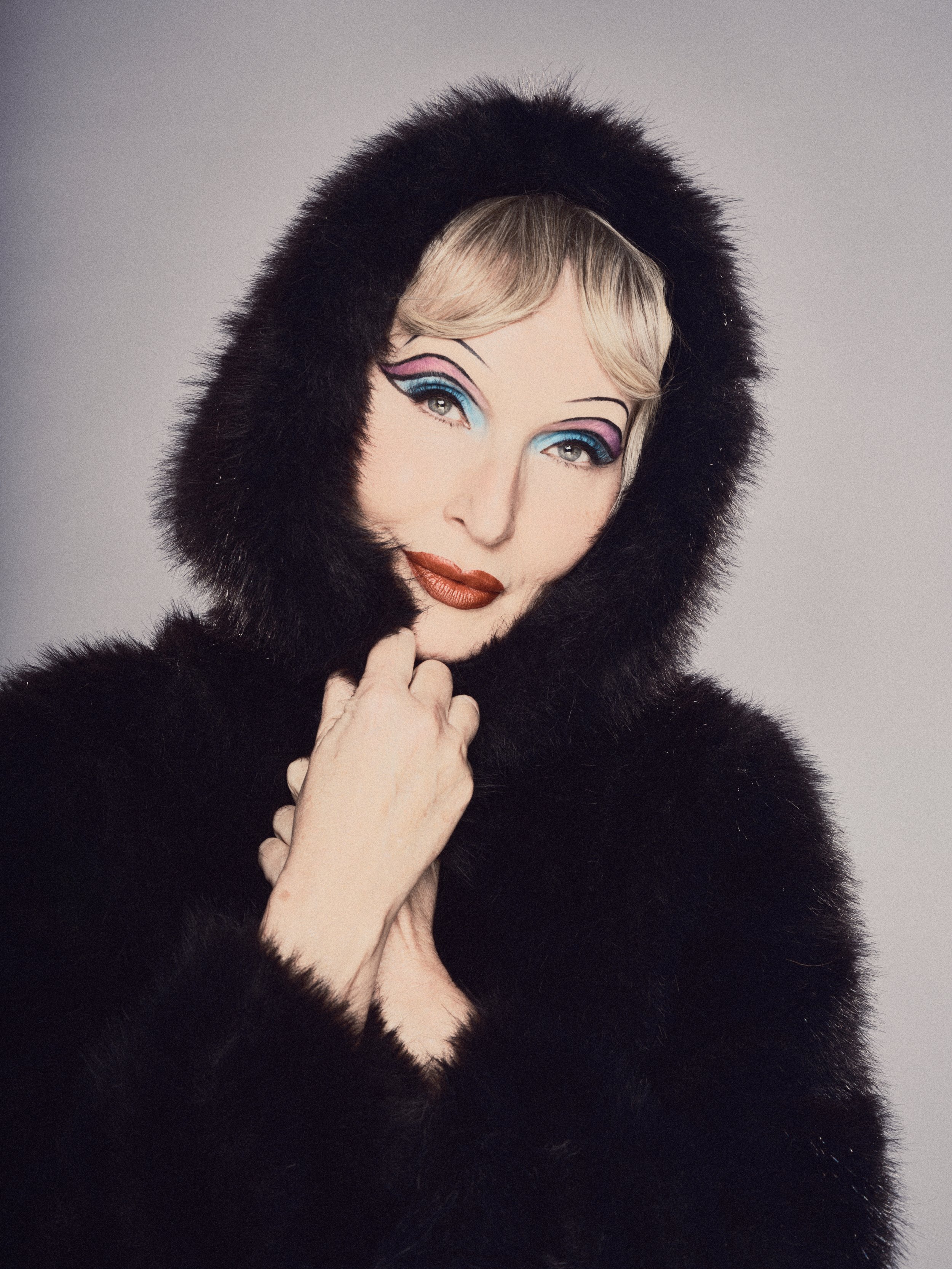 Portrait of a woman with styled blonde hair and colorful makeup, wearing a black fur hood and coat, against a plain background.