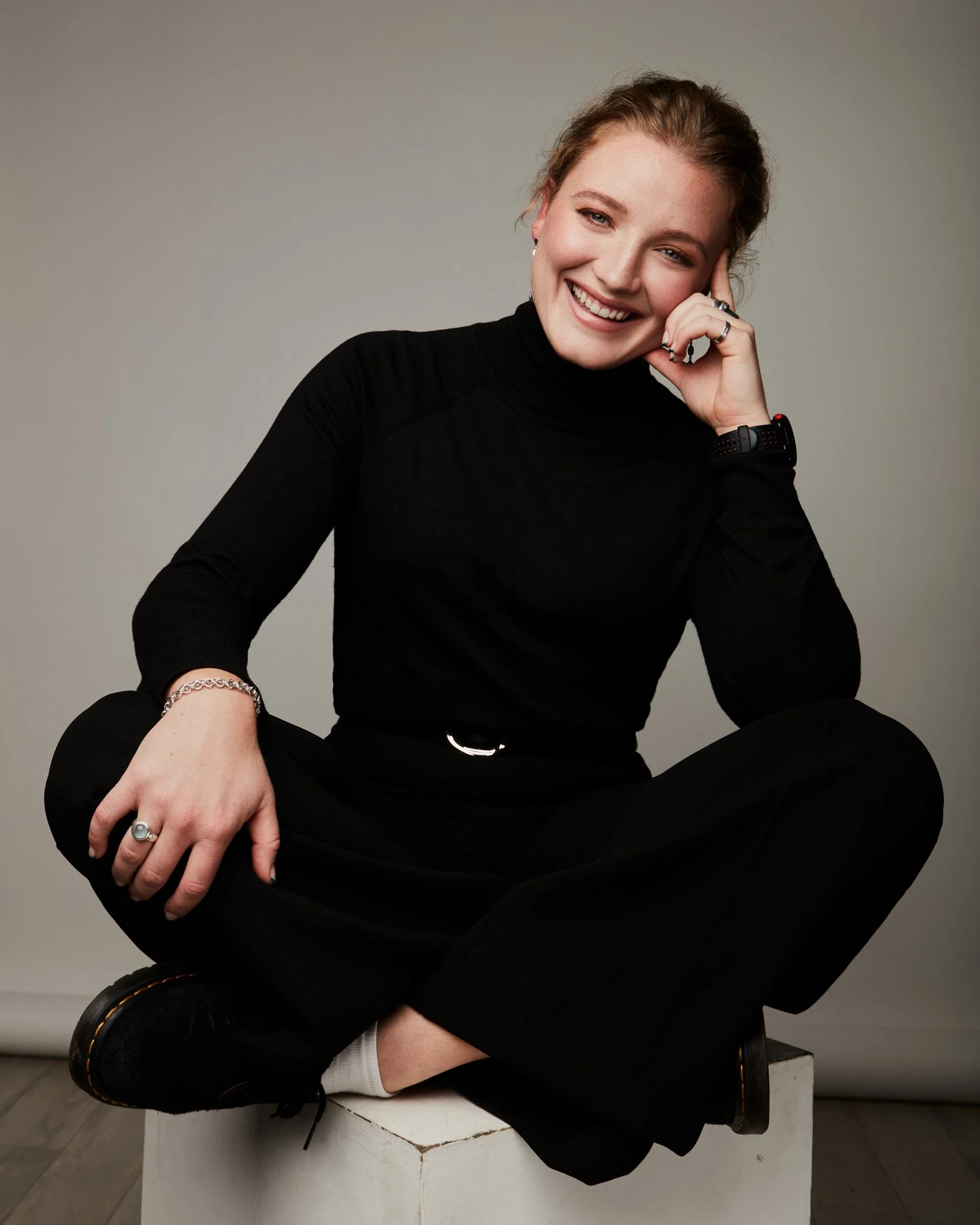 A young woman with red hair, wearing a black turtleneck top, jewelry, and a smartwatch, sitting cross-legged on a white surface and smiling at the camera with her hand resting on her face.