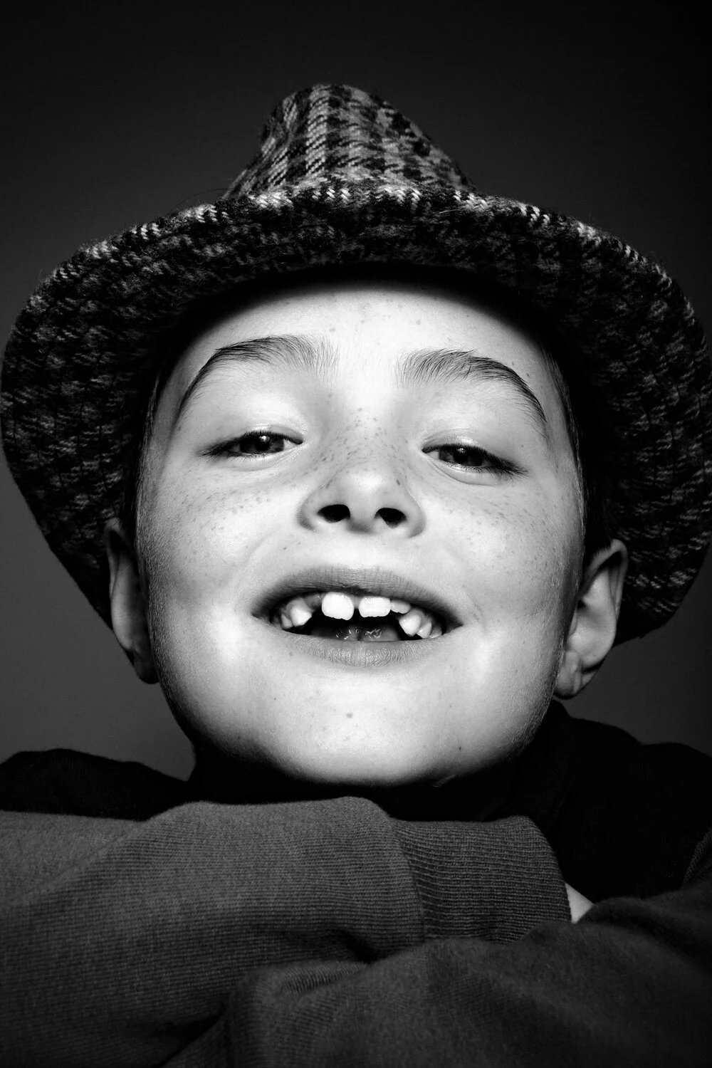 A smiling young boy with missing front teeth wearing a knit hat and dark clothing, looking directly at the camera, in black and white.