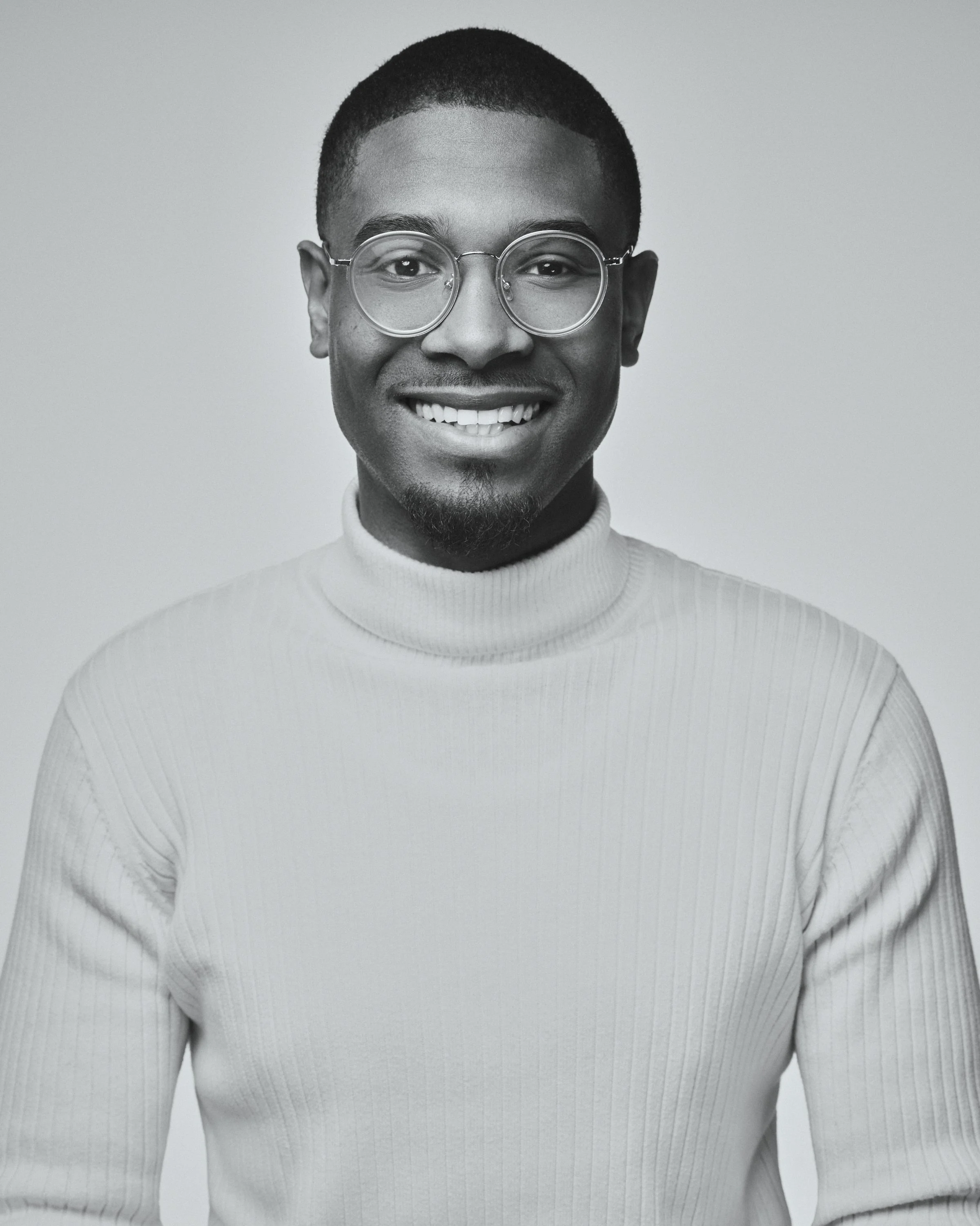 A black-and-white portrait of a smiling man wearing glasses and a ribbed turtleneck sweater, against a plain background.