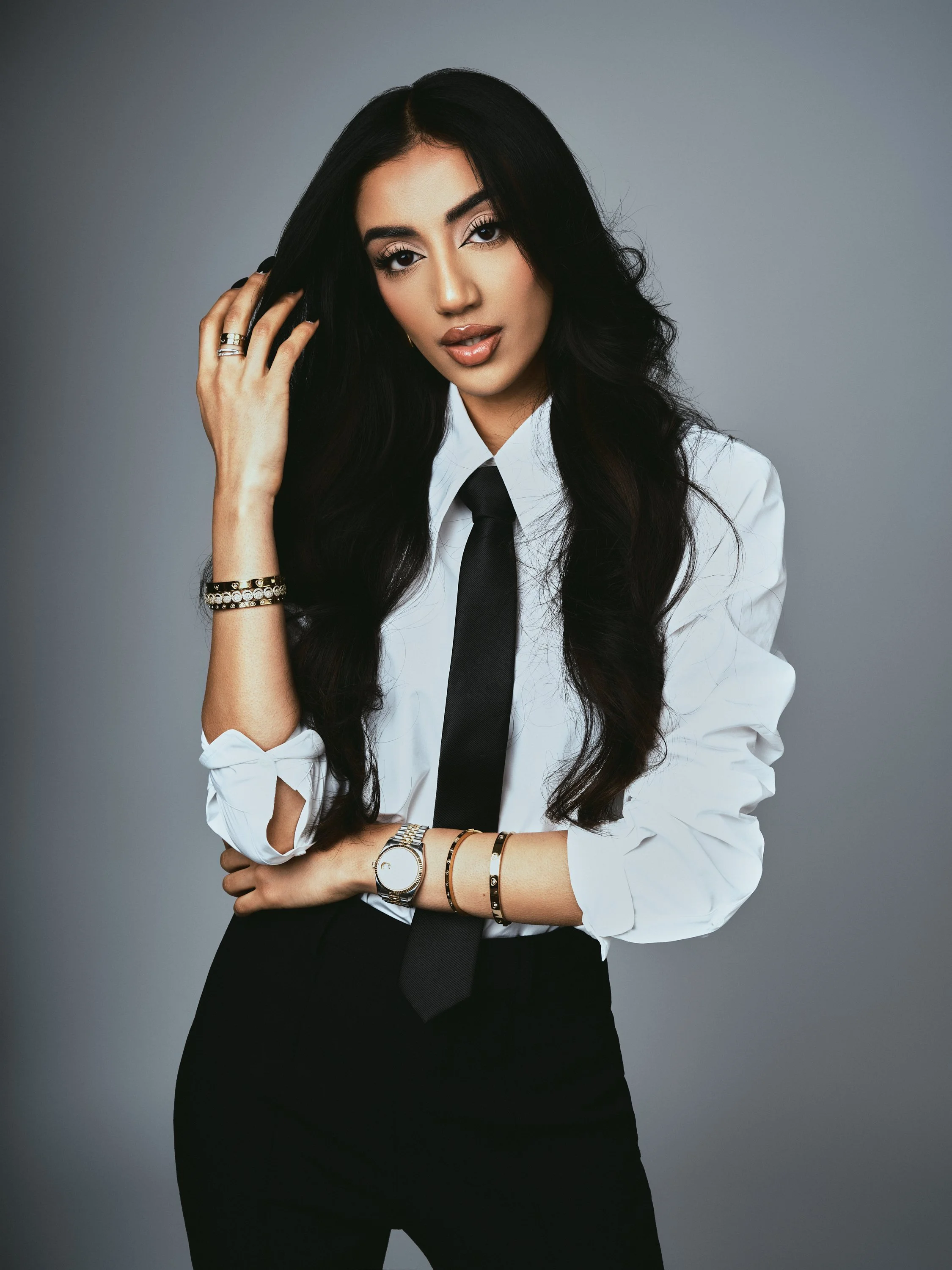 A woman with long black hair, wearing a white shirt, black tie, and black pants, posing against a gray background.