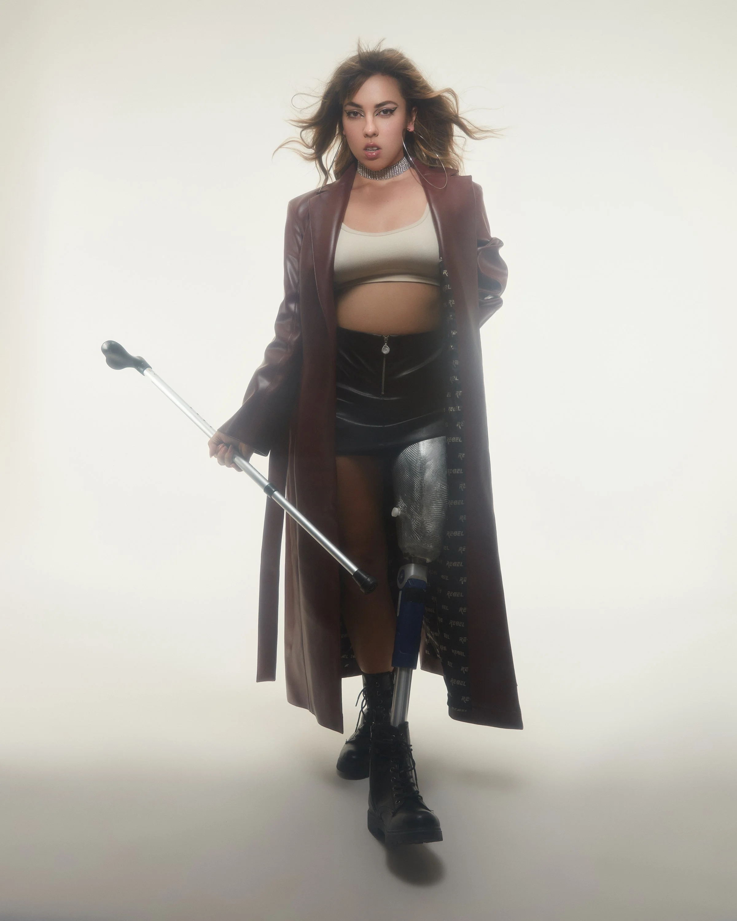 A woman with wavy hair wearing a brown leather jacket, a white crop top, a black leather skirt, black boots, and holding a cane, standing in front of a plain background.