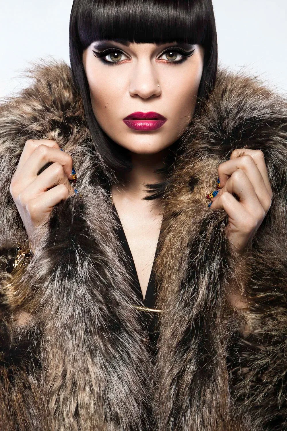 A woman with dark, straight hair and bangs wearing a fur coat, with bold makeup including dark eyeliner and deep red lipstick.