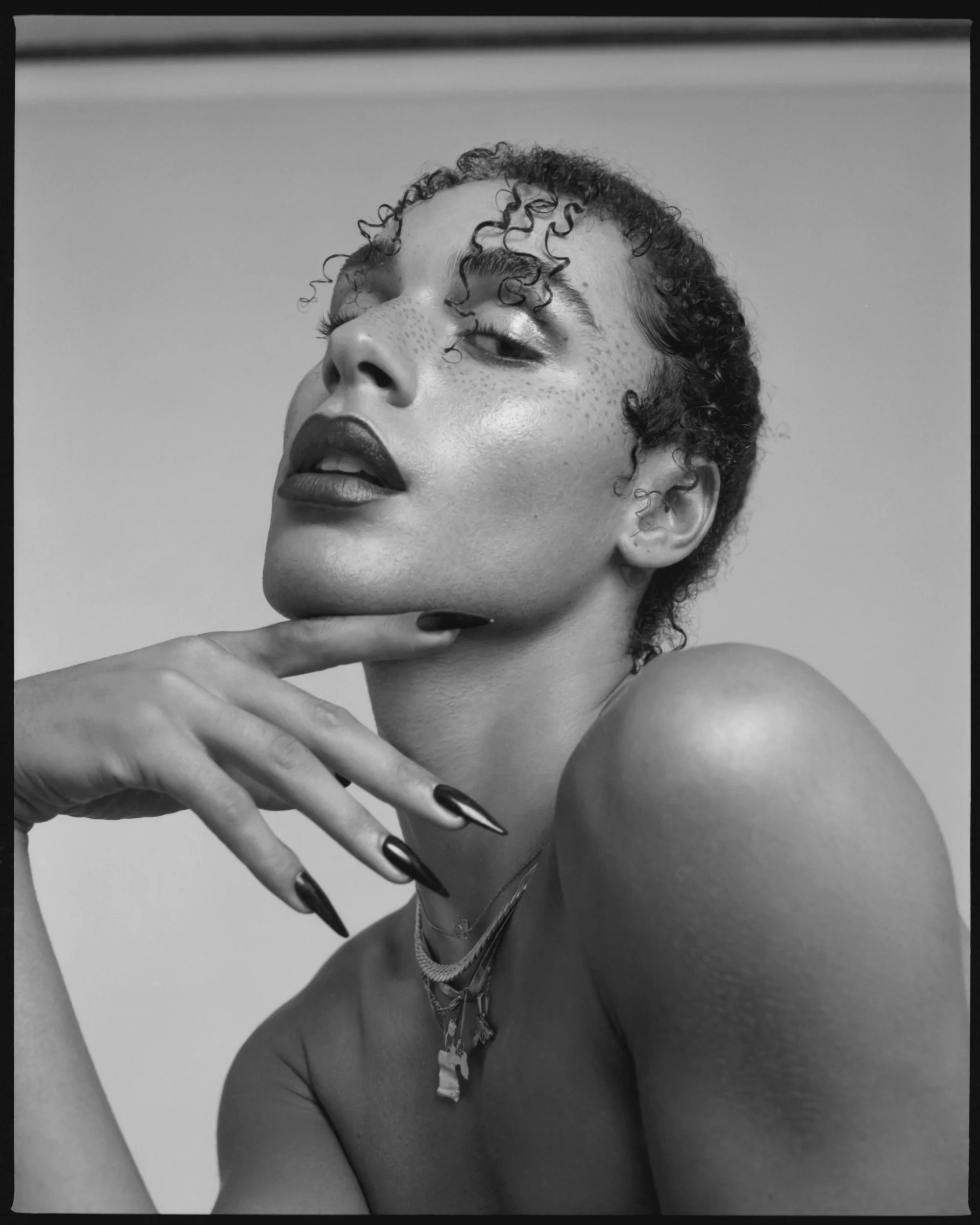 Black and white portrait of a woman with short curly hair, dark lipstick, and multiple necklaces, posing with her hand near her chin and looking slightly upward.