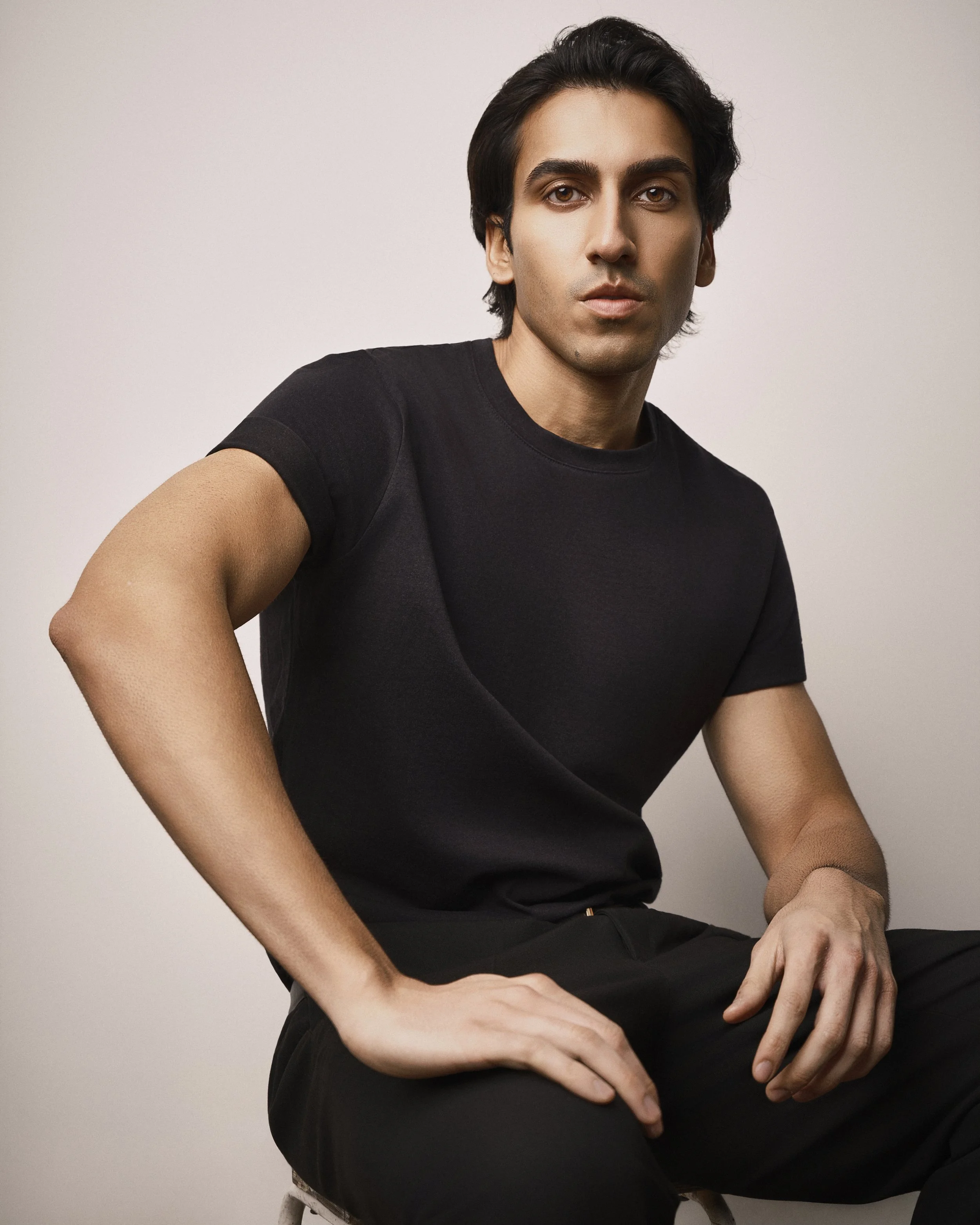 A young man with dark hair and fair skin wearing a black t-shirt, seated with his arms resting on his lap, against a plain light-colored background.