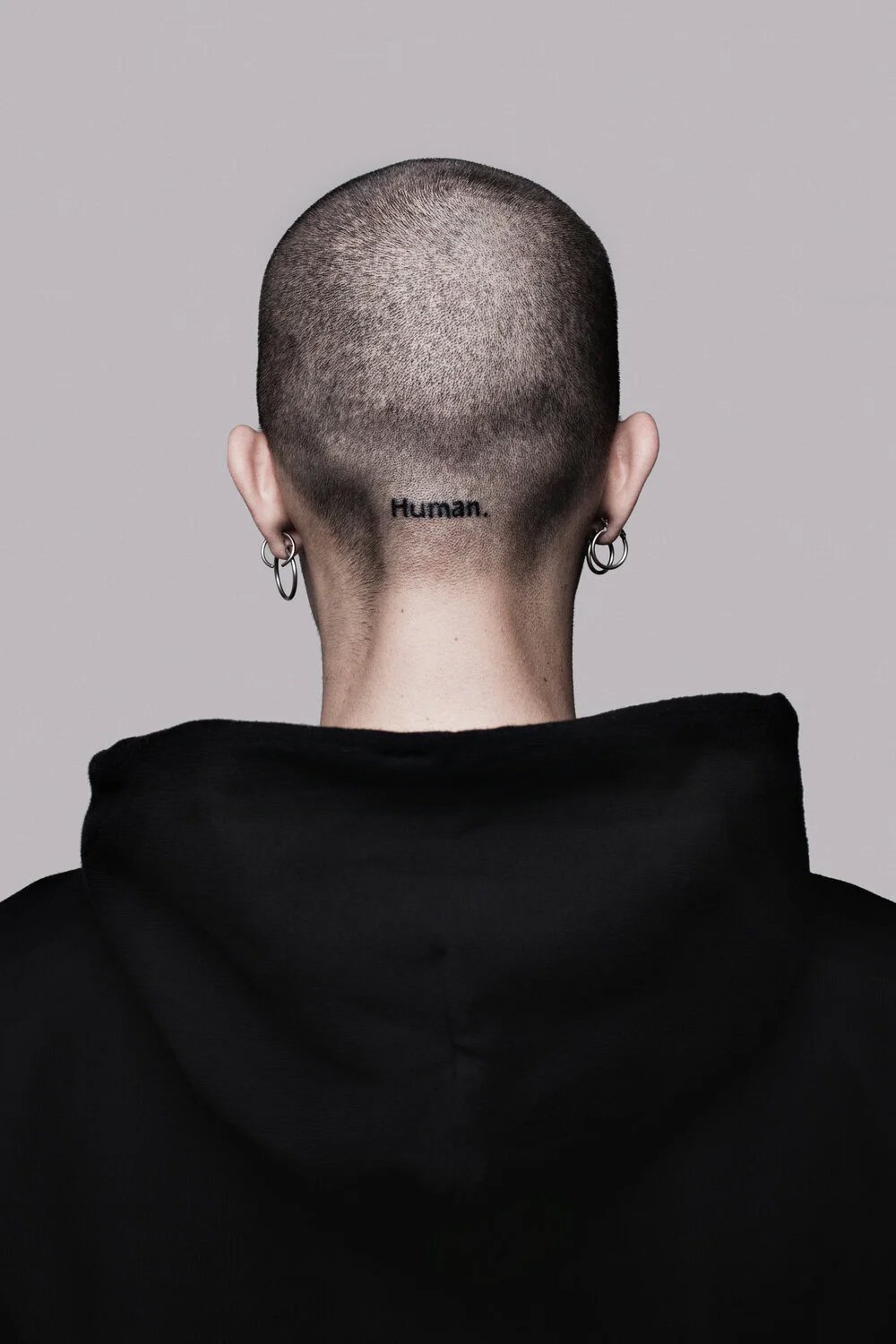 Back of a person with a shaved head and multiple earrings, wearing a black hoodie, with the word "Human" tattooed on the back of their neck.