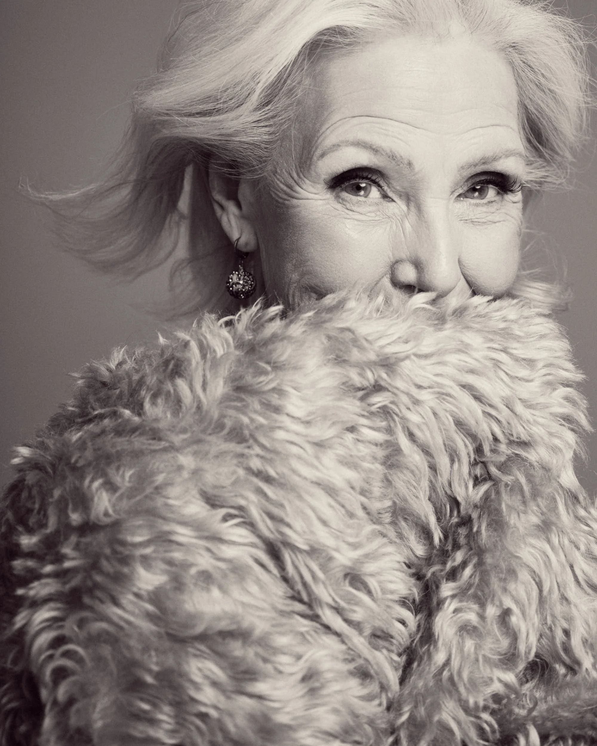 Close-up of an elderly woman with white hair, wearing earrings, partially covered with a furry blanket, looking at the camera.