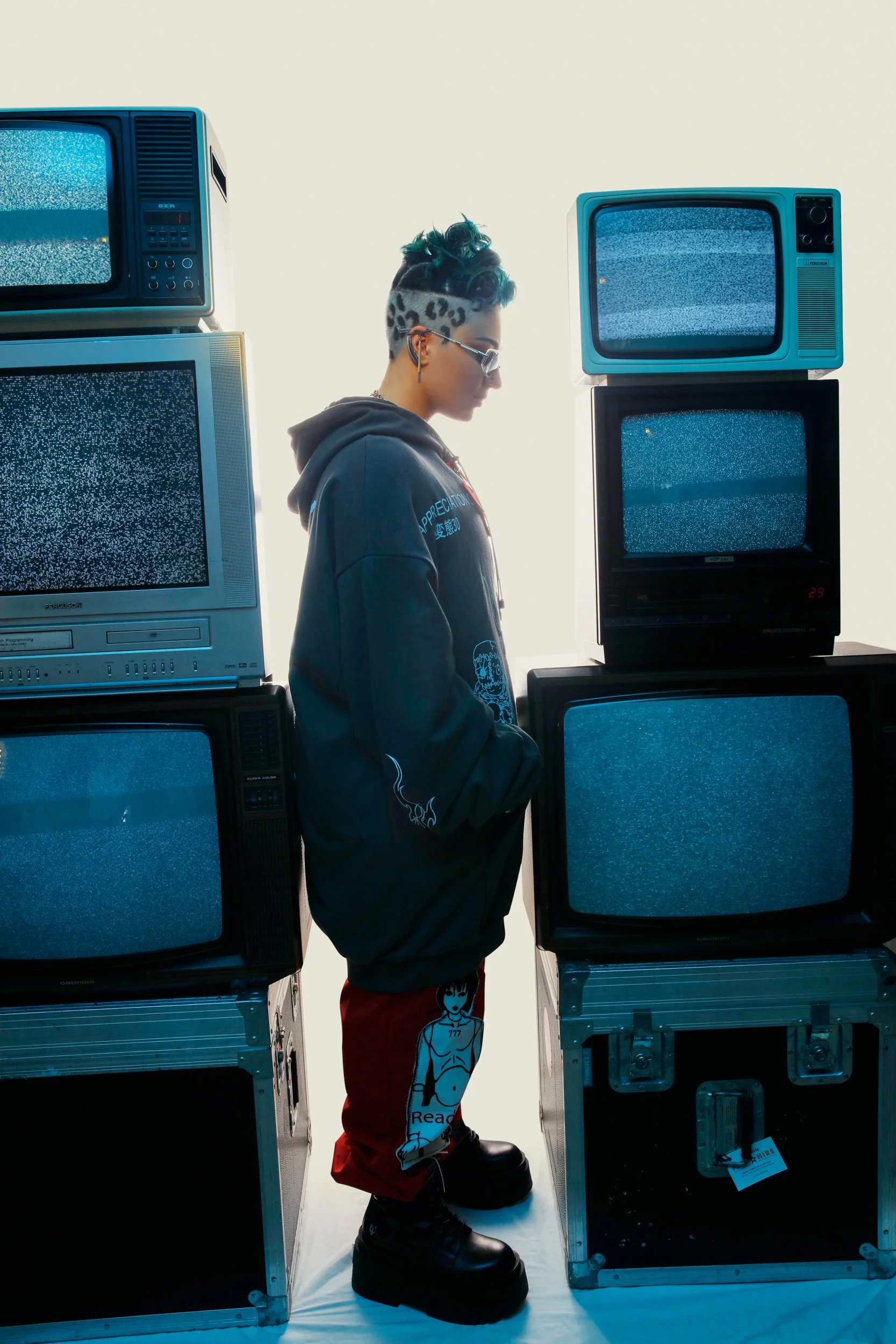 A young person with short mohawk hairstyle, leopard print pattern, and sunglasses stands among a stack of vintage television sets displaying static.