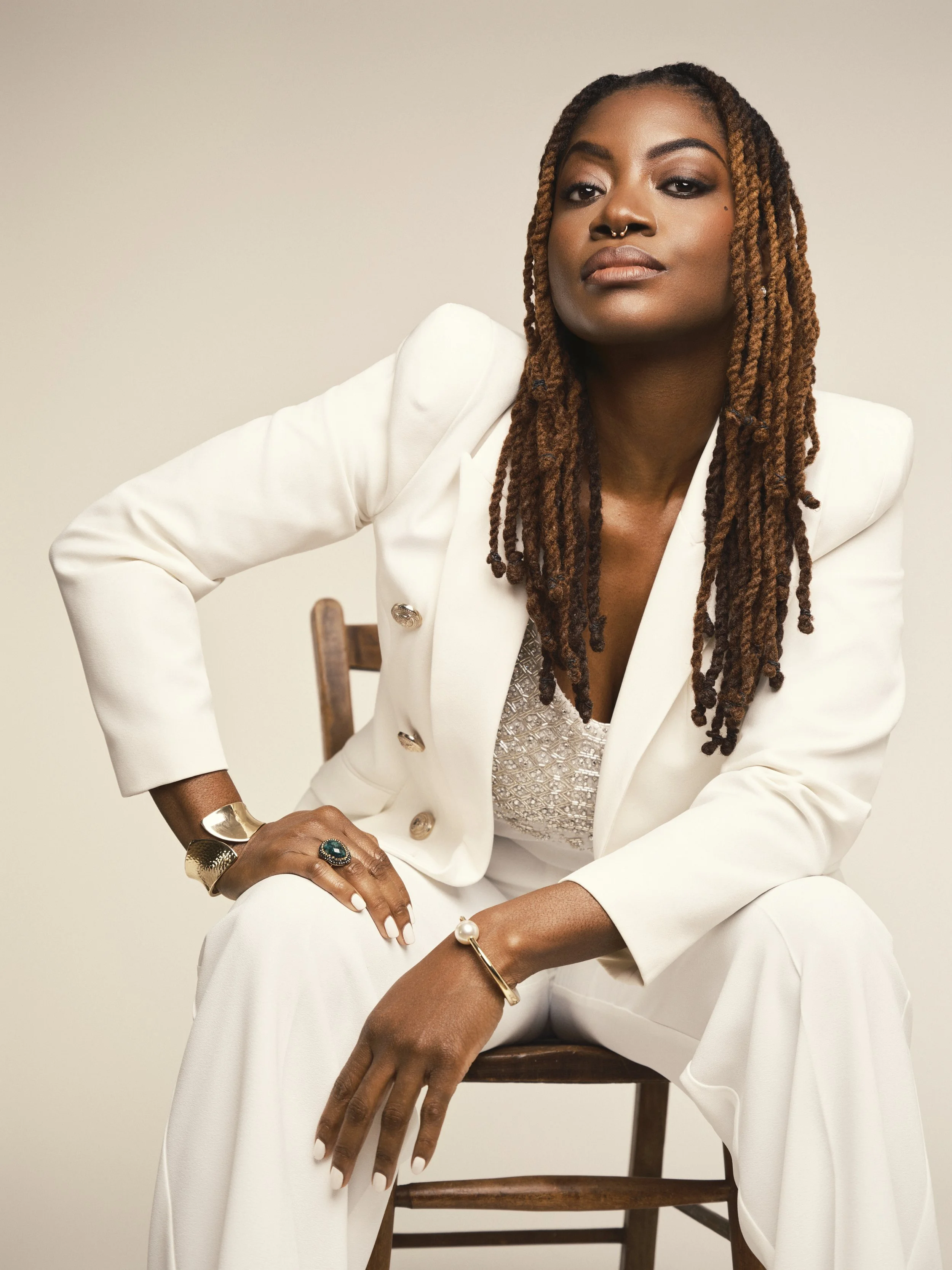 Portrait of a confident woman with dreadlocks, wearing a white suit with gold buttons, sitting on a wooden chair against a plain beige background.