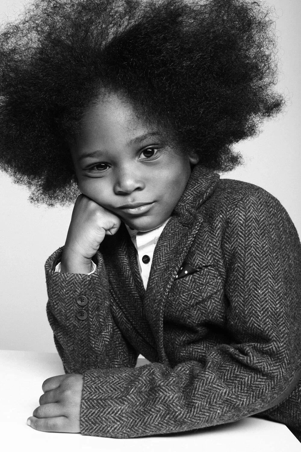 A young girl with voluminous natural hair, resting her head on her hand, wearing a textured blazer and a collared shirt, in a portrait shot.