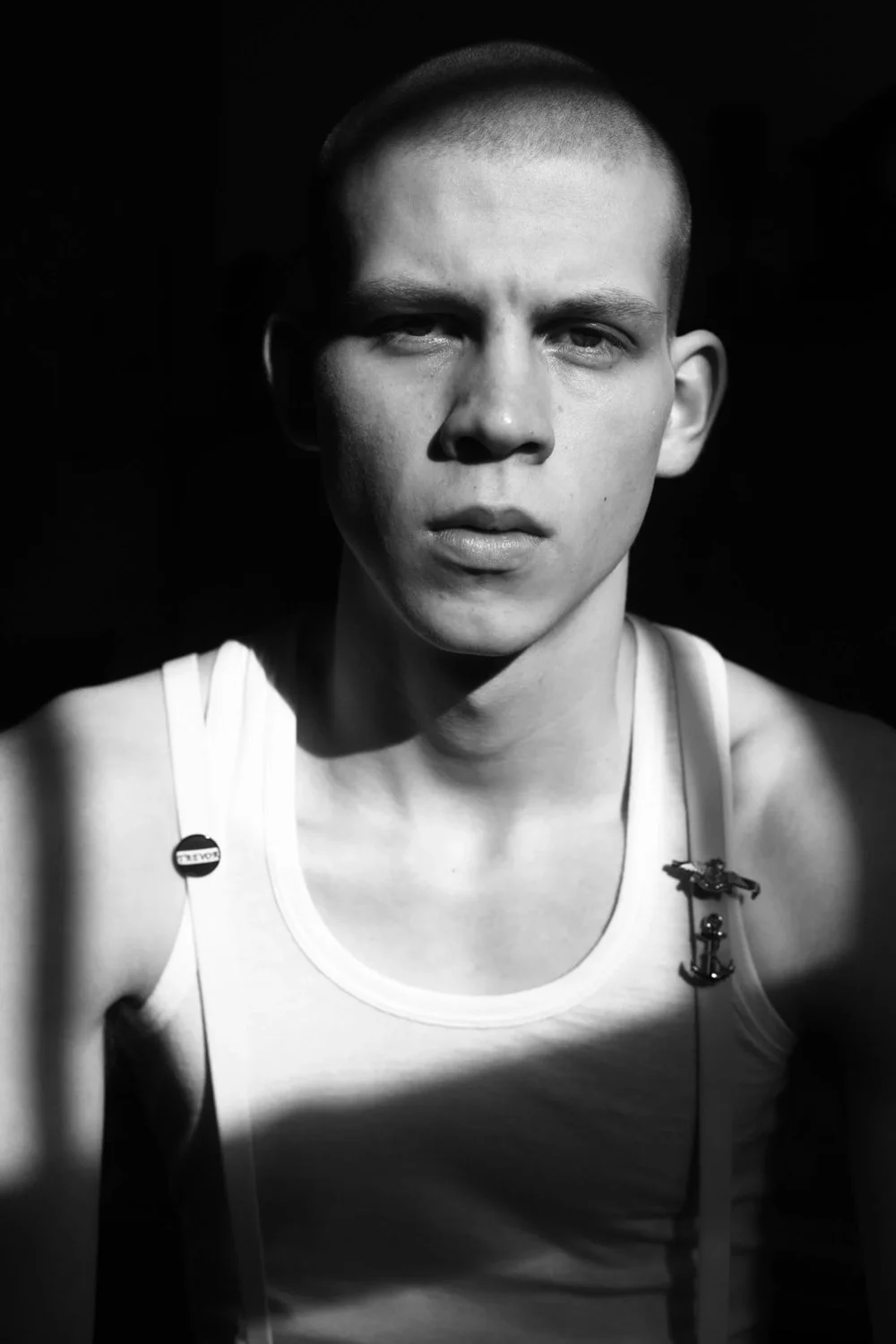Black and white close-up of a young man with a buzz cut hairstyle, wearing a white tank top with a small button on the strap and a small decorative anchor pin.
