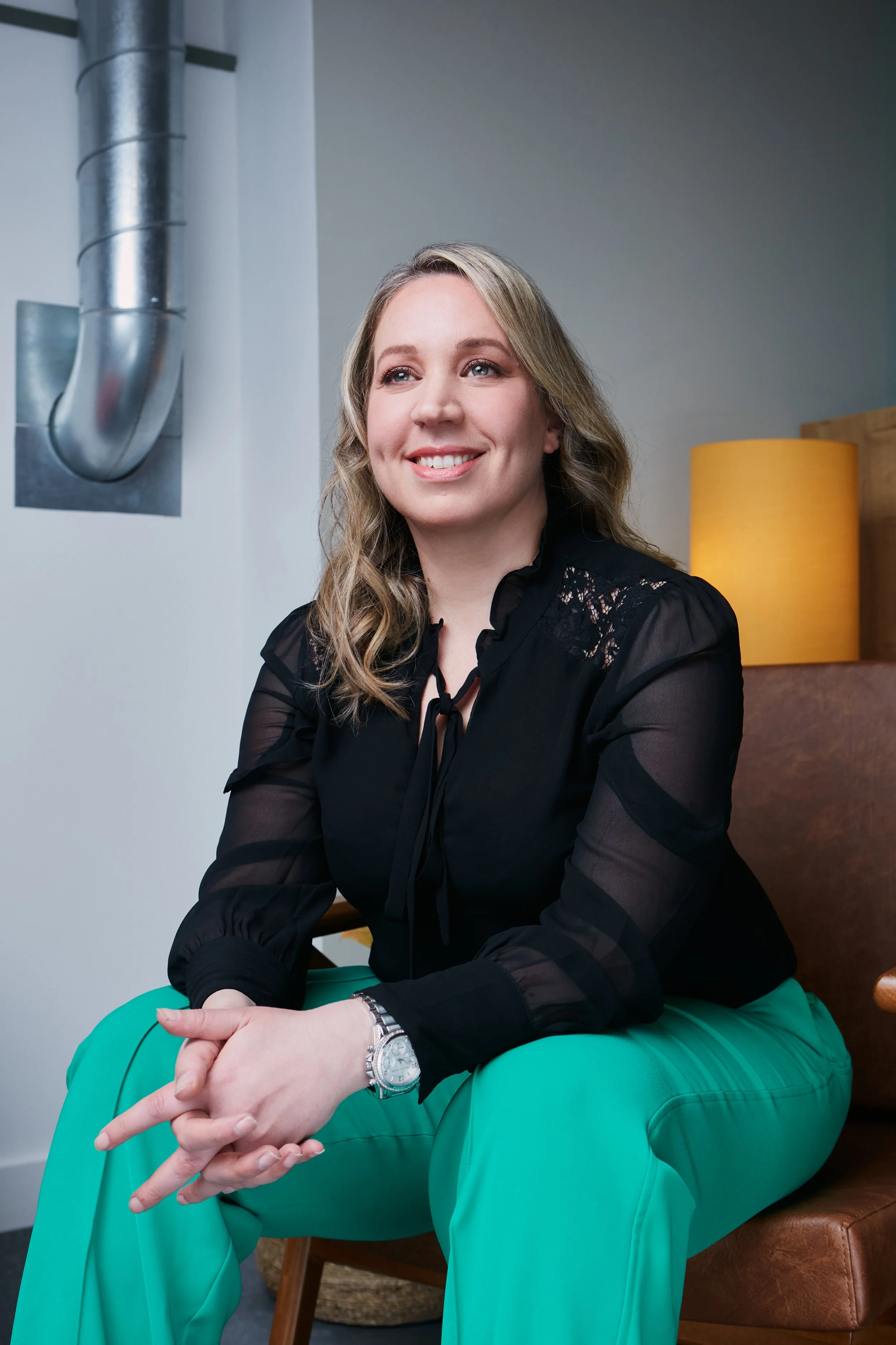 A woman with blonde hair smiling, wearing a black top and bright green pants, sitting on a green chair in a modern indoor setting with a metal duct, a yellow lamp, and wooden furniture in the background.