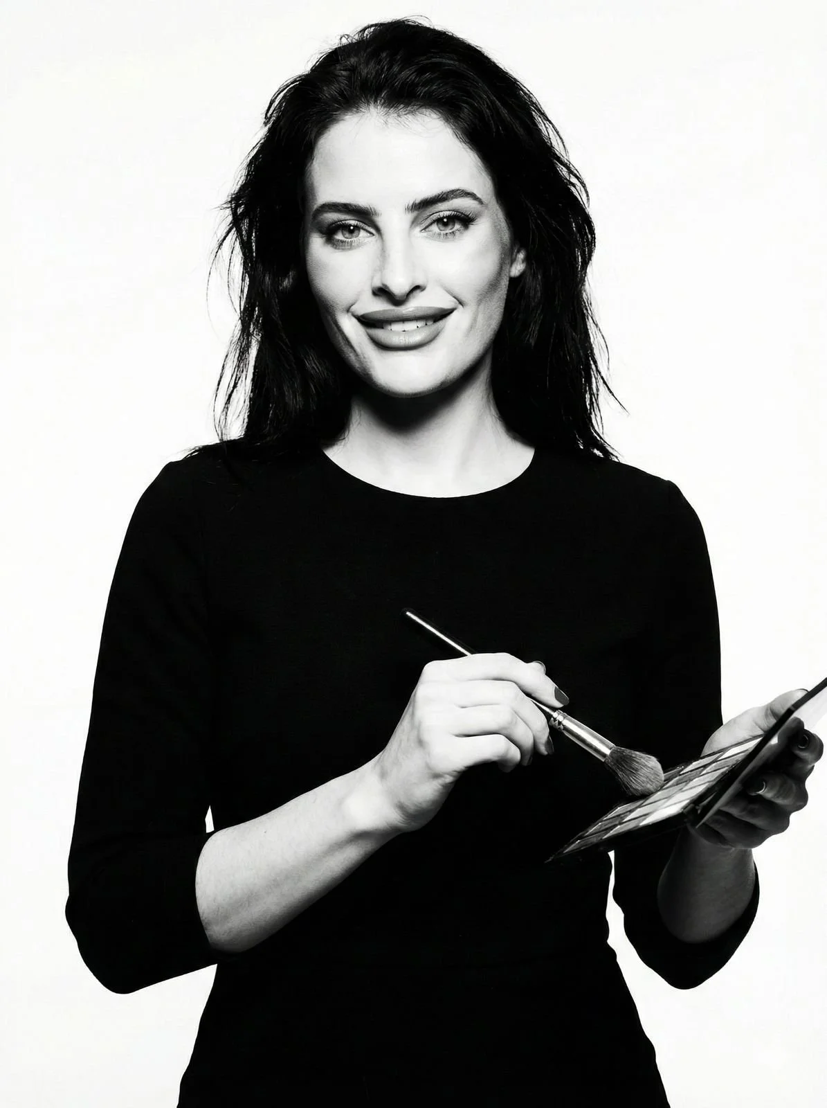 Black and white portrait of a smiling woman with long dark hair, bright eyes, and makeup, wearing a necklace and earrings.