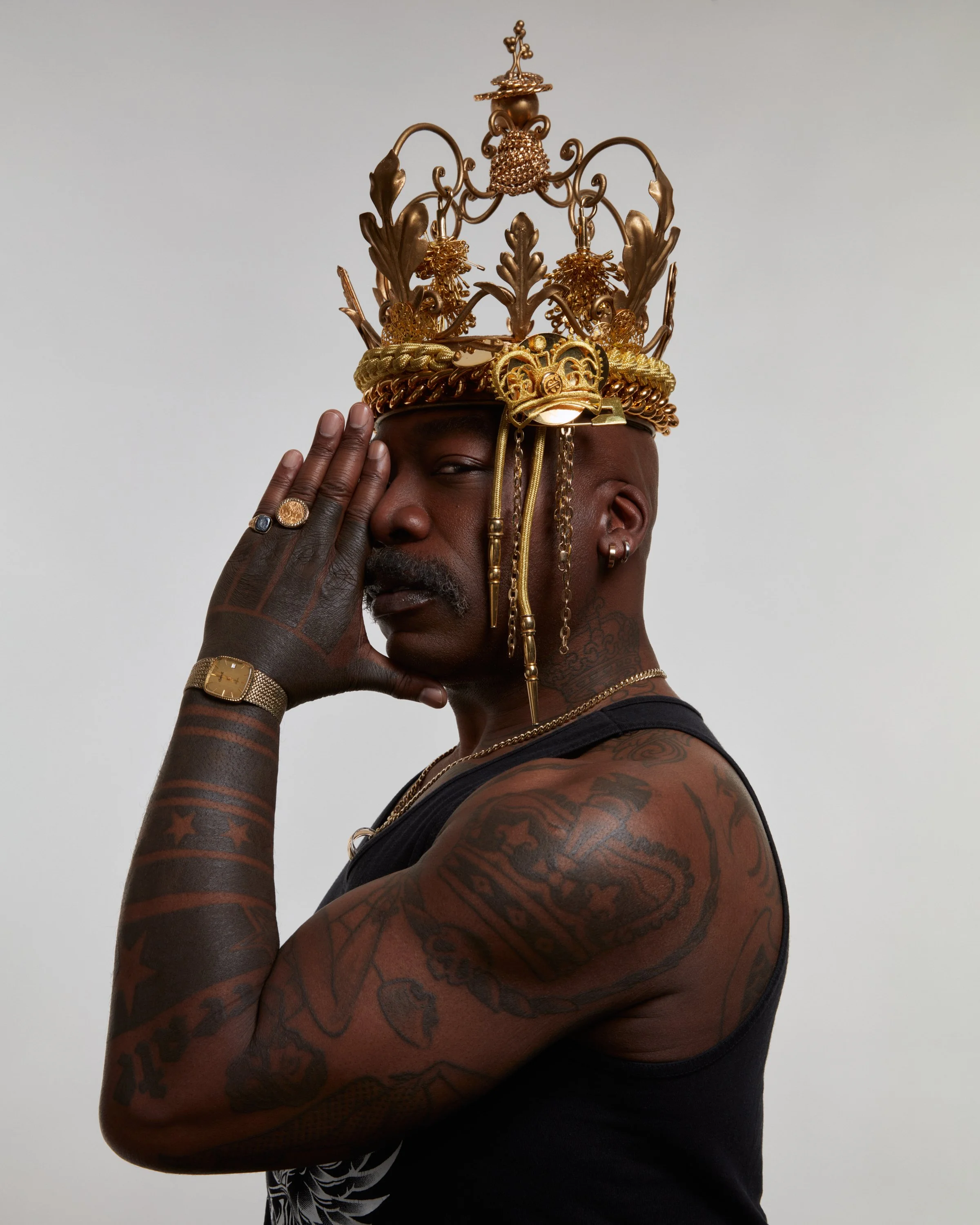 A man with tattoos wearing a black sleeveless shirt, gold jewelry, and a large ornate gold crown, posing with his hand near his face against a plain background.