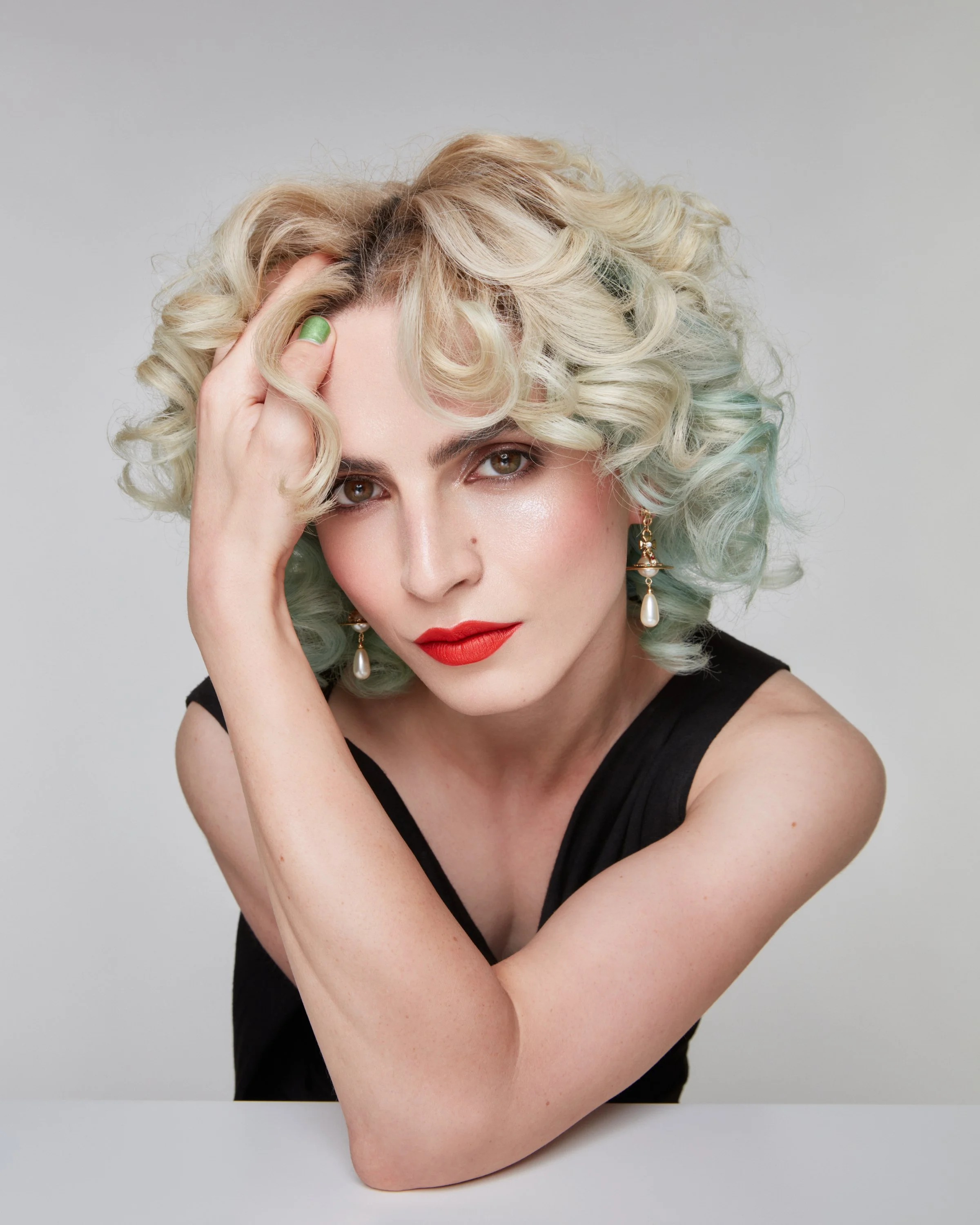 A woman with platinum blonde, curly hair, red lipstick, and wearing pearl earrings poses against a plain background.