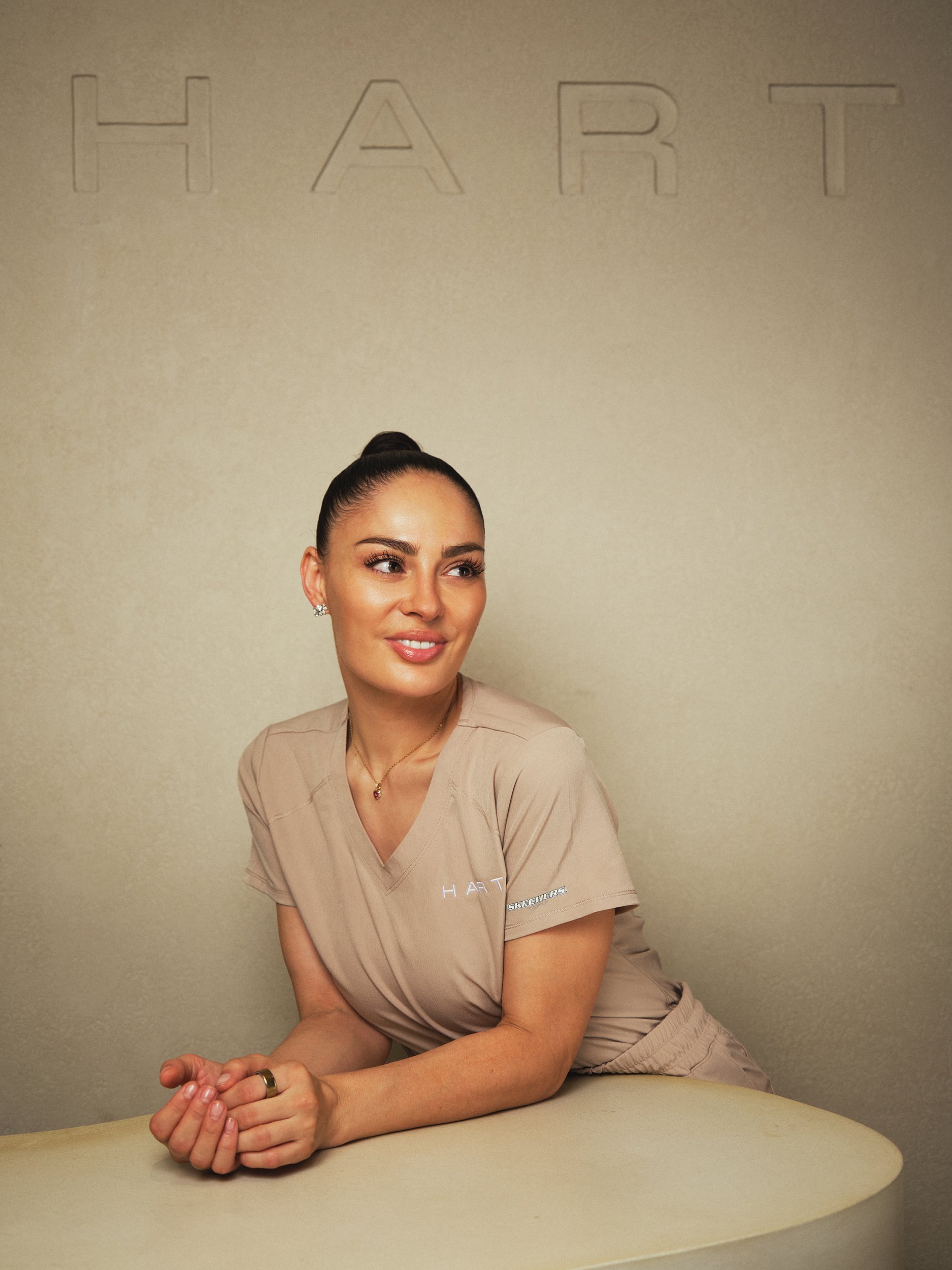 A woman with dark hair in a bun, wearing tan medical scrubs, seated at a table with her hands clasped, smiling slightly, against a beige wall with the word 'HART' faintly visible at the top.