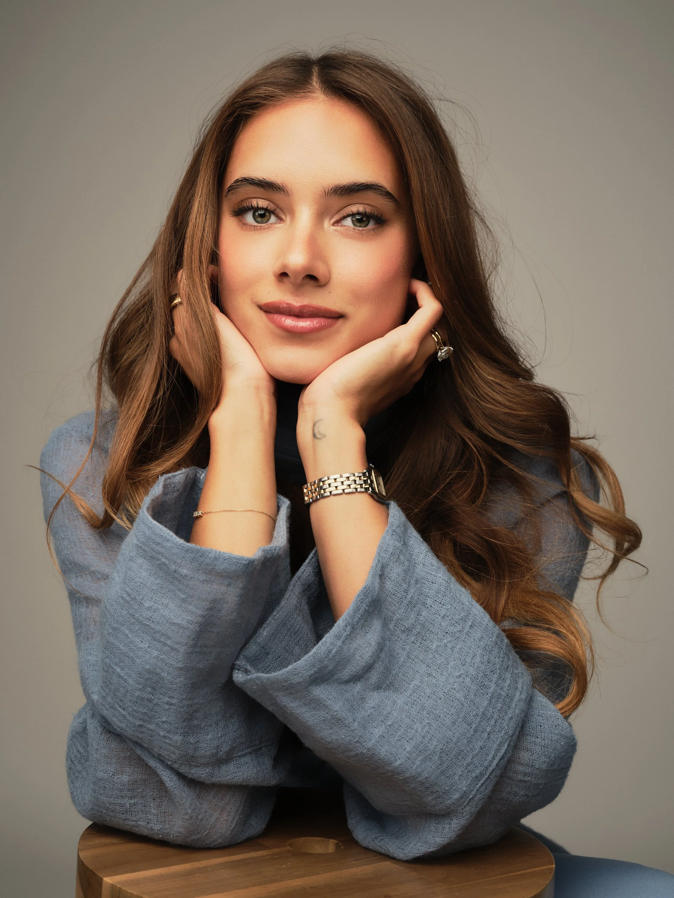A portrait of Ambar Lee, A young woman with long brown hair, wearing a blue blazer, rests her face in her hands and smiles at the camera against a neutral background.