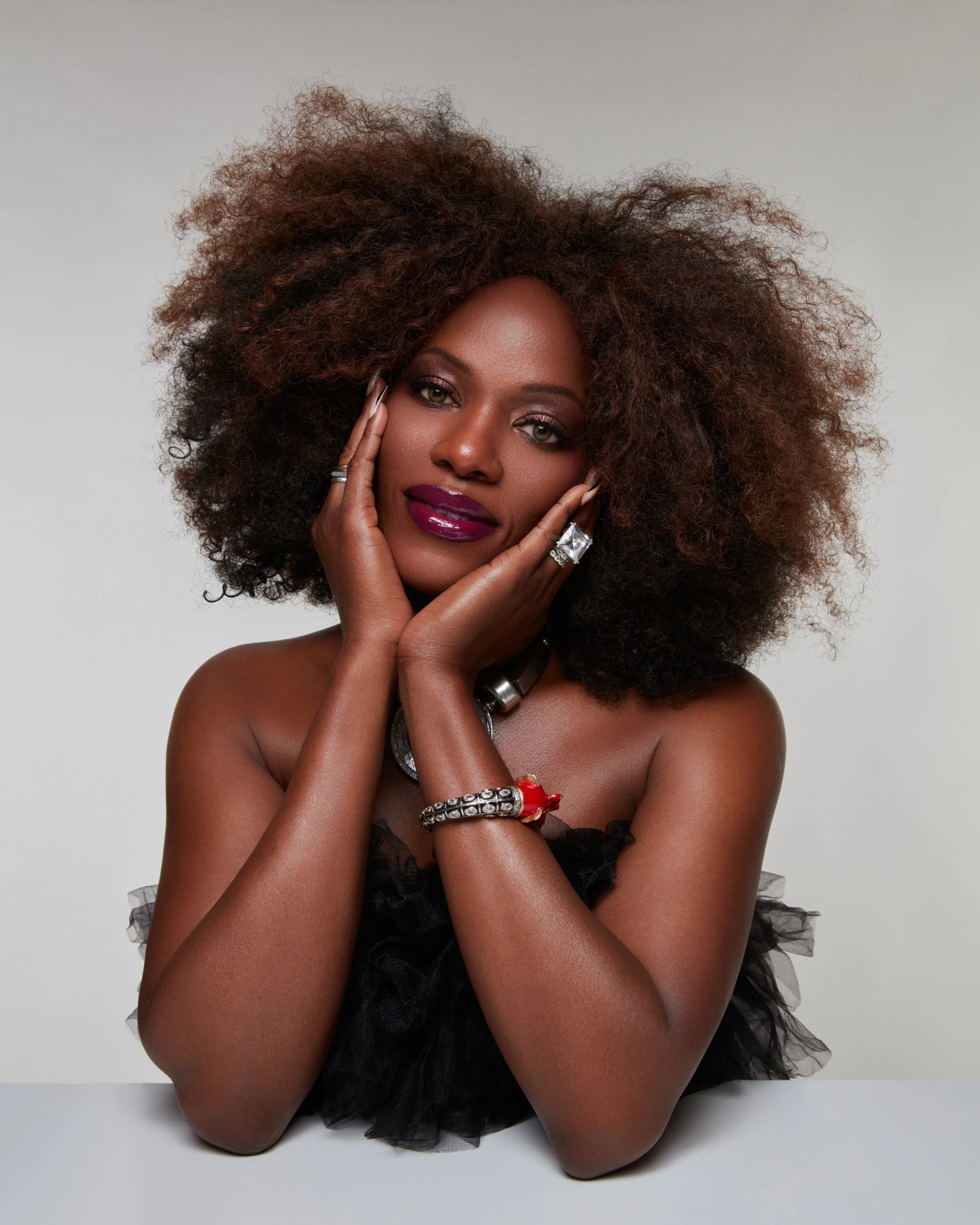 A woman with dark skin and voluminous curly hair poses with her head resting on her hands, wearing jewelry and makeup, against a neutral background.