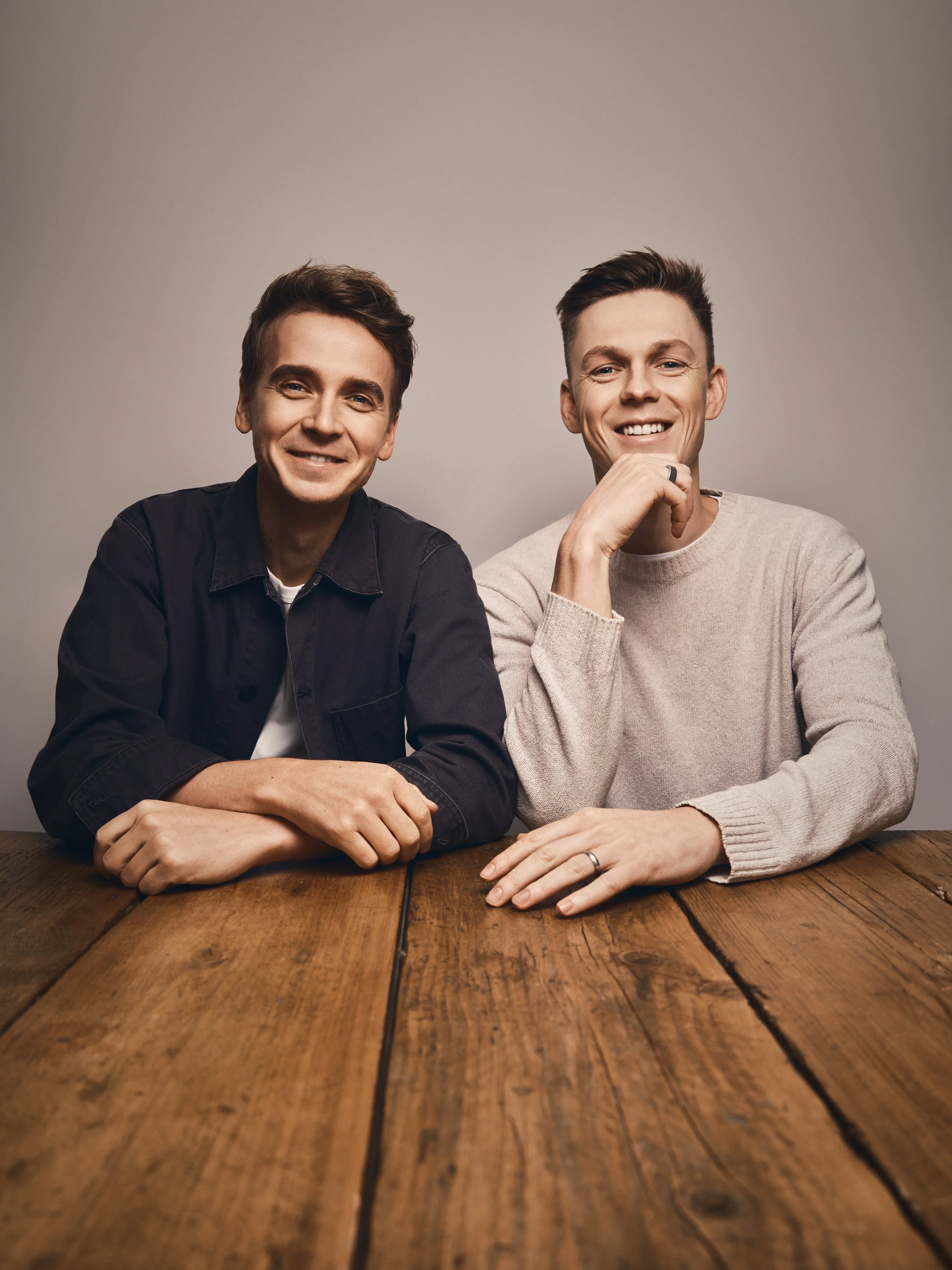 Joe Sugg & Caspar Lee sitting at a wooden table, smiling at the camera, against a plain background.