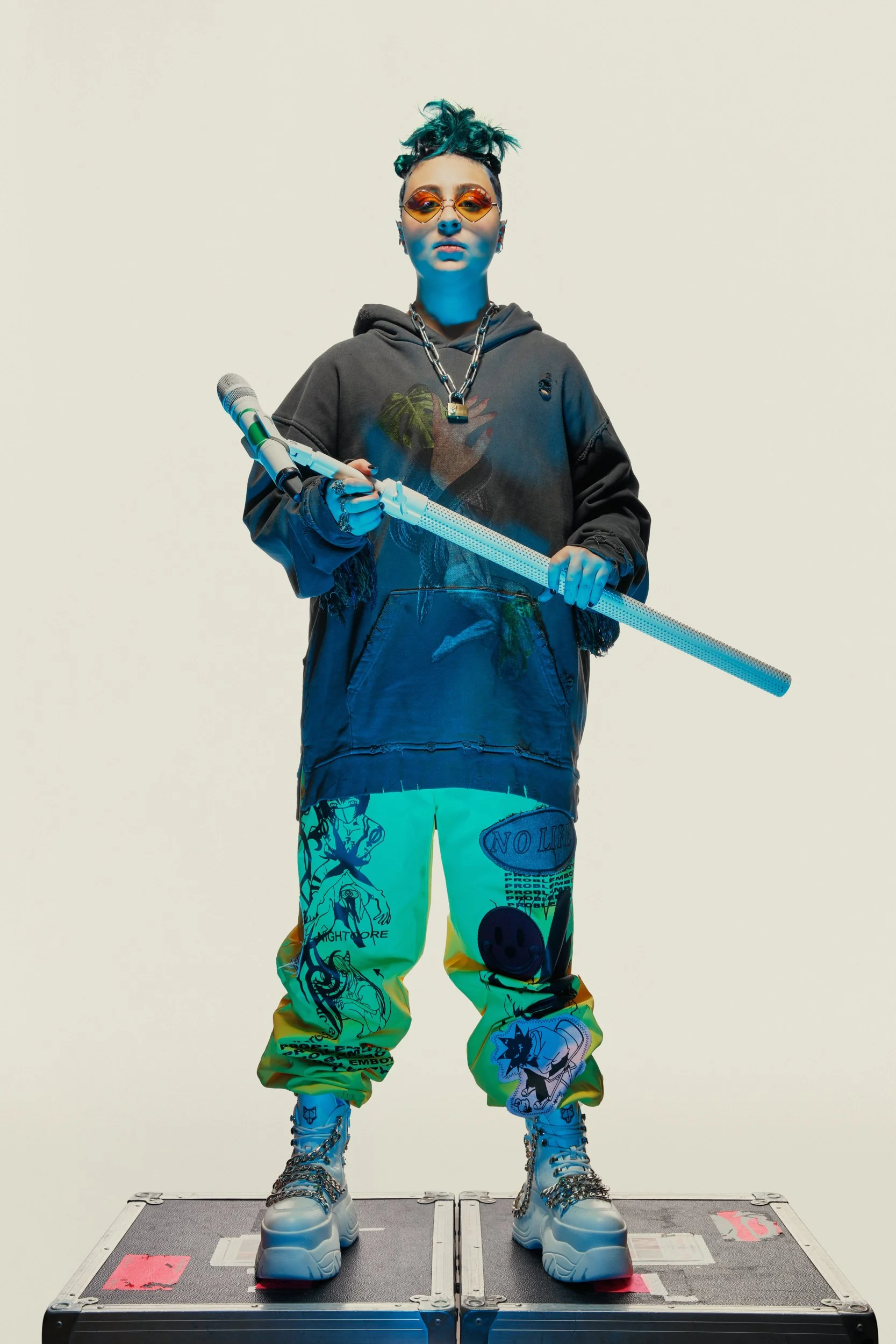Fashionably dressed person standing on a gray platform, holding a ray gun prop, wearing colorful graphic pants, chunky boots, a hoodie, sunglasses, and jewelry, against a plain light background.