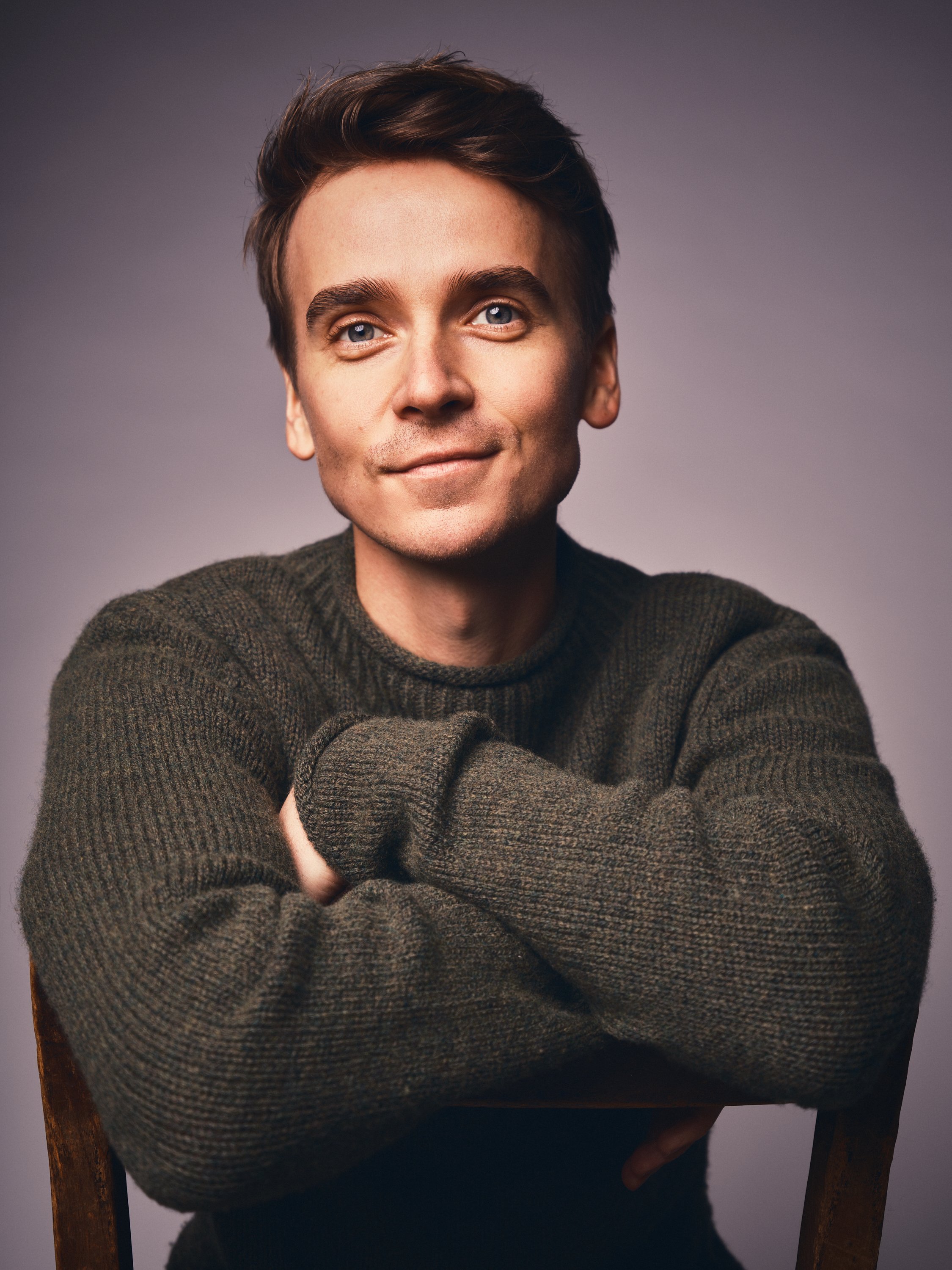 Portrait of a young man with brown hair and blue eyes, wearing a dark green knit sweater, sitting with arms crossed, against a neutral background.