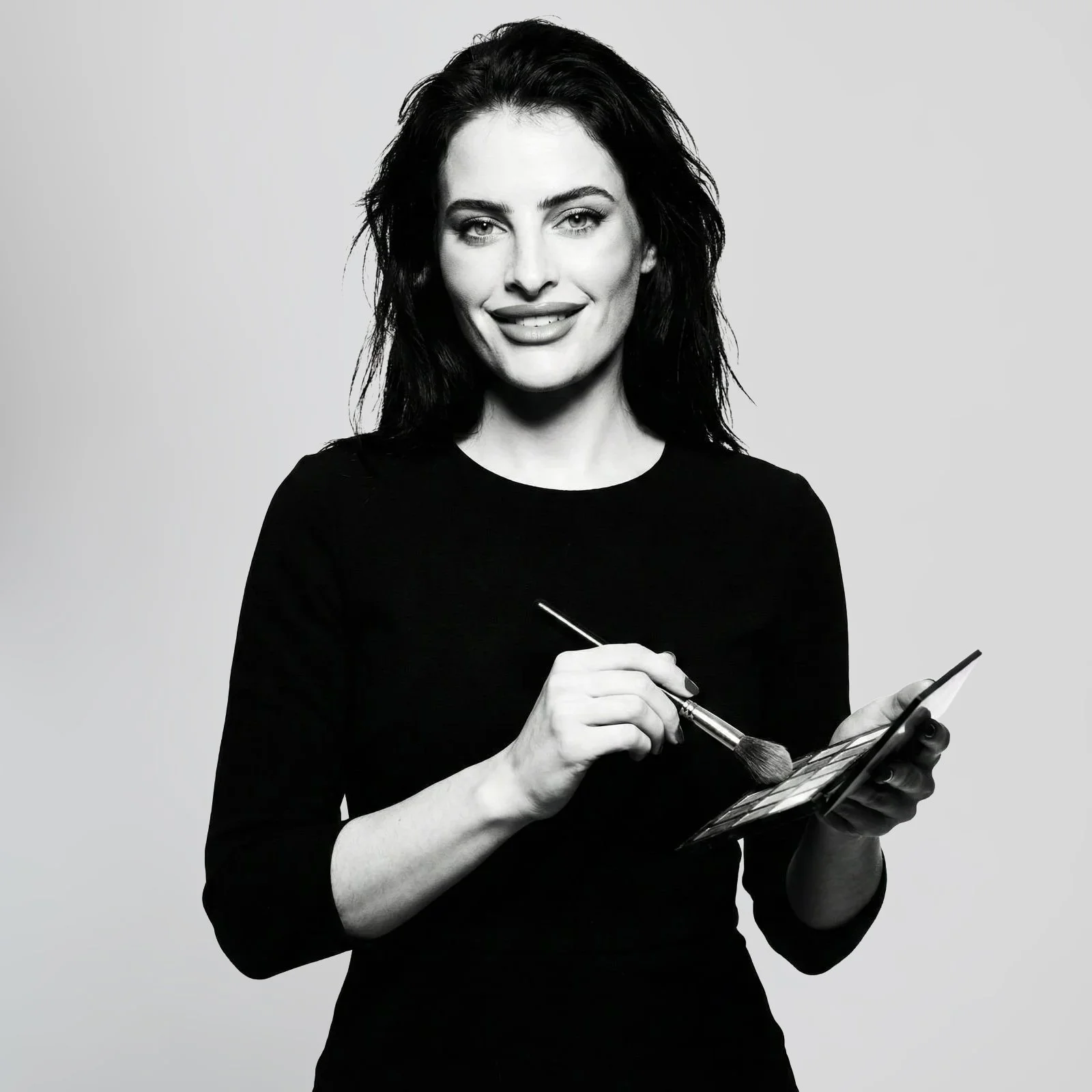 Black and white portrait of a smiling woman with long dark hair, bright eyes, and makeup, wearing a necklace and earrings.