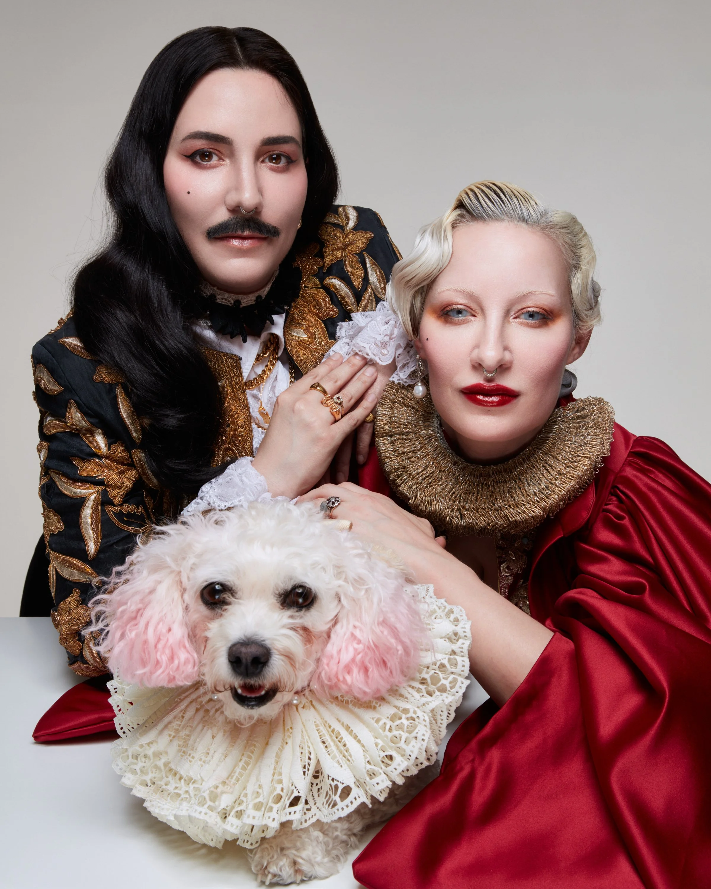 Two women dressed in elaborate, vintage-style clothing with makeup and jewelry, and a white poodle dog with pink-dyed ears and a lace collar, all against a plain background.