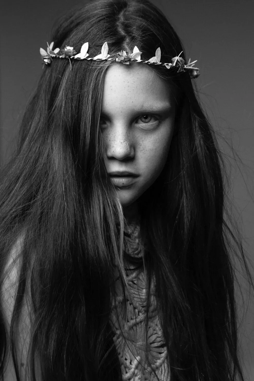 A black and white portrait of a young woman with long hair, wearing a floral crown and a textured top, staring intensely at the camera.