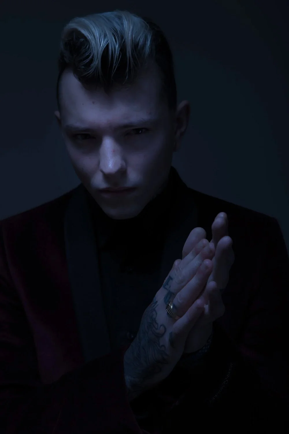 A young man with pale skin and short, platinum blonde hair with dark roots, looking intensely at the camera, with hands pressed together in a prayer-like pose, wearing a dark jacket in a dimly lit setting.