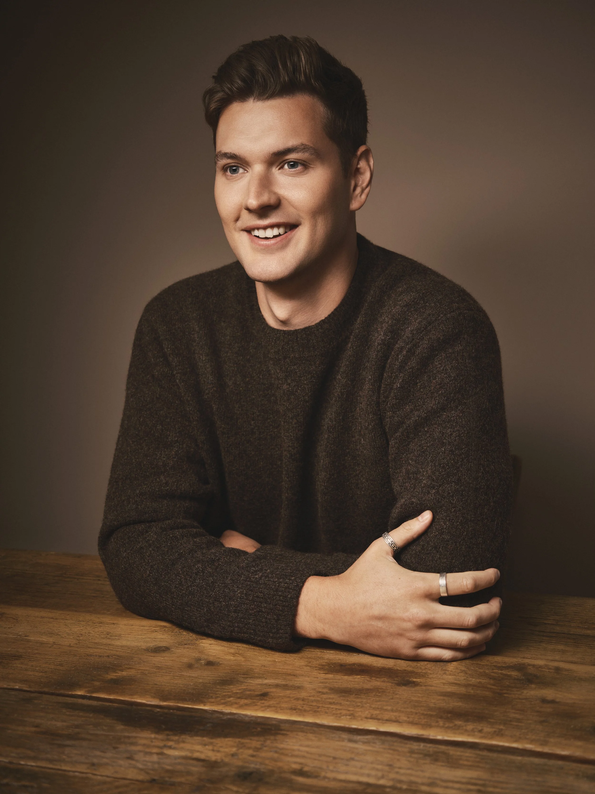 A young man with short brown hair, blue eyes, wearing a dark brown sweater, sitting at a wooden table with a neutral dark background.