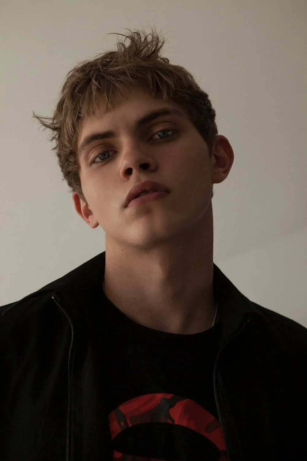 A young man with light skin, tousled light brown hair, and piercing blue eyes looking directly at the camera. He's wearing a black jacket and a black shirt with a red graphic design.