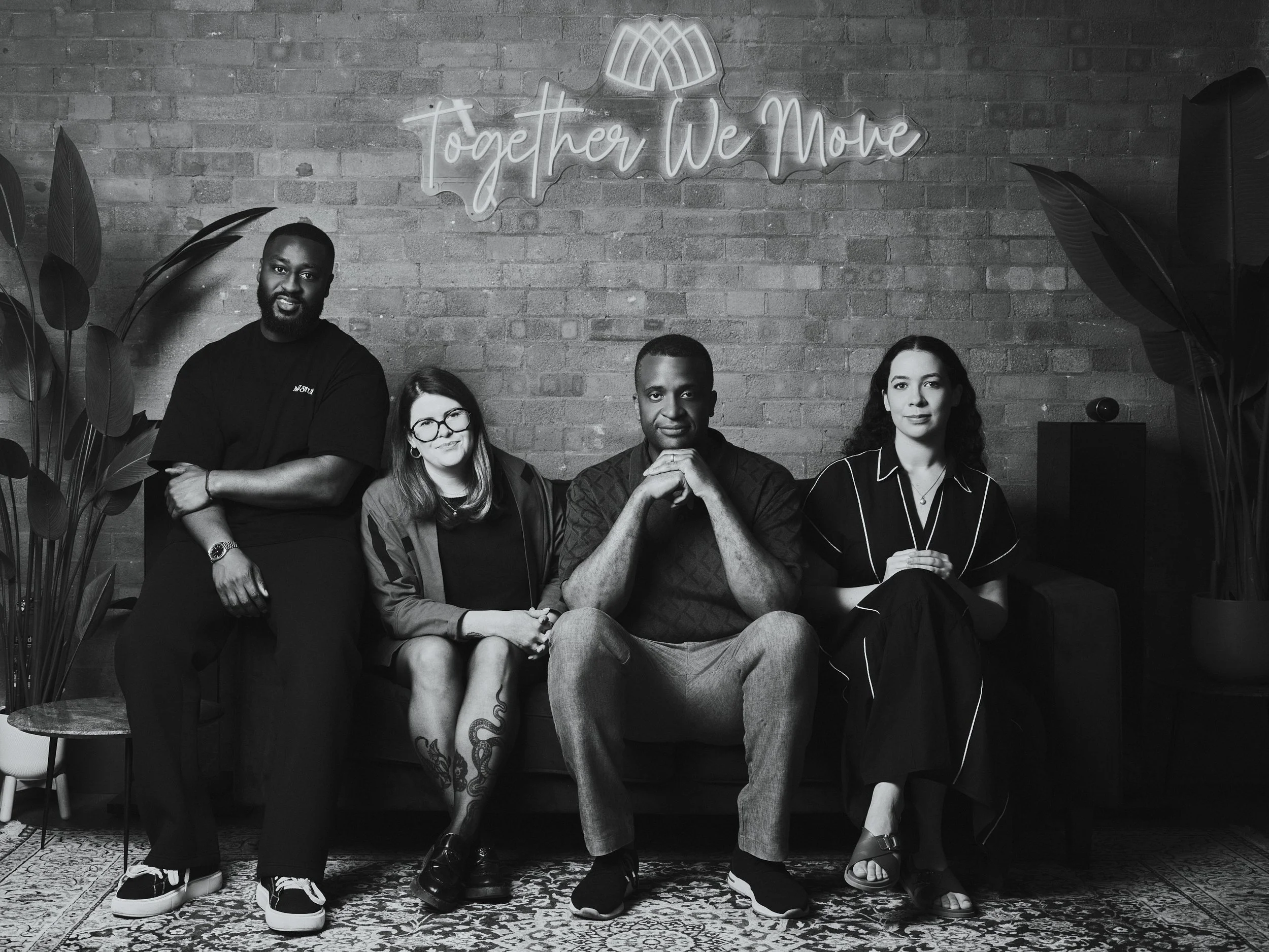 Four diverse people sitting on a sofa with a brick wall in the background, a neon sign reading 'Together We Move,' and large plants on either side.