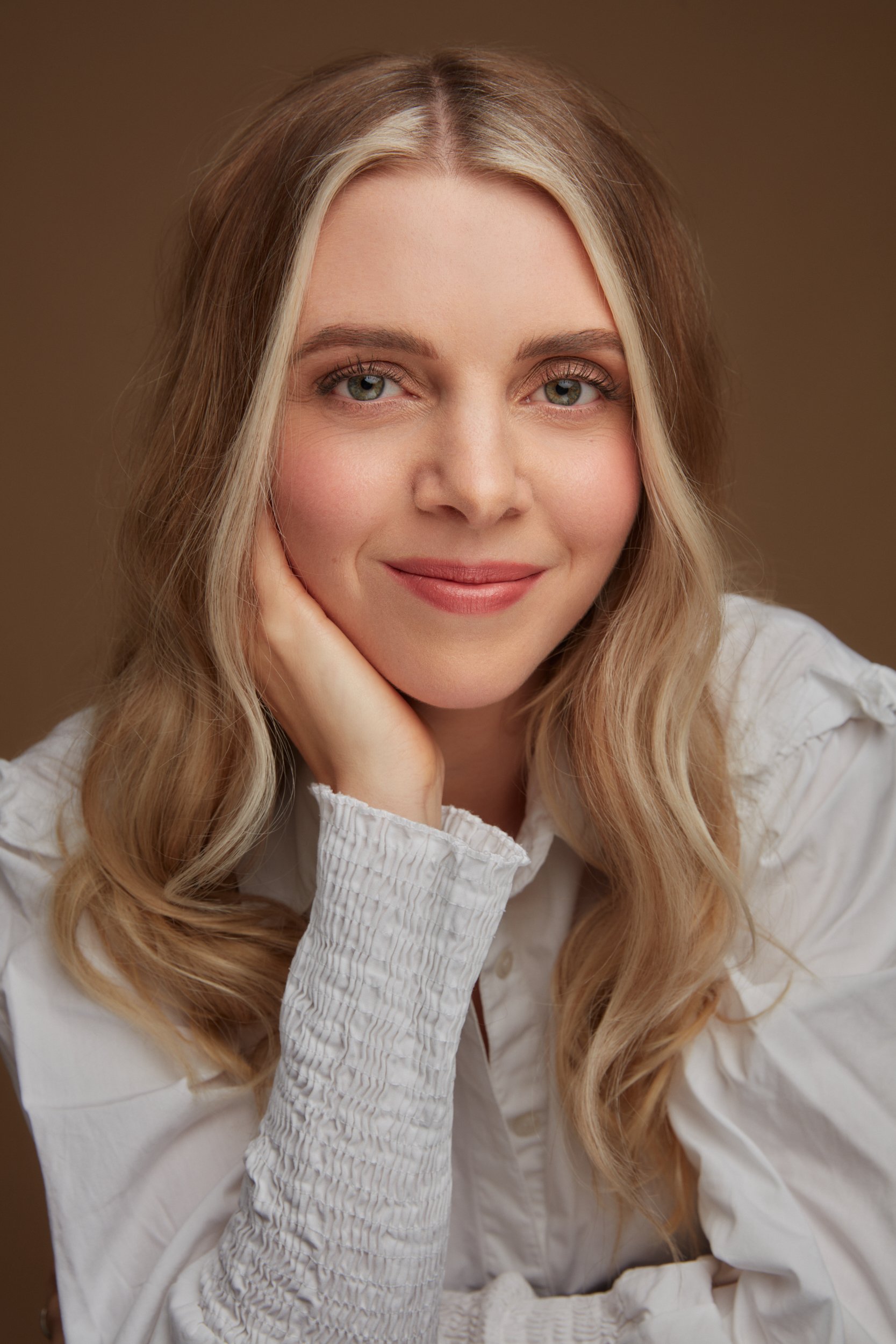 Close-up of a woman with long, blonde hair and blue eyes, smiling softly and resting her chin on her hand, wearing a white, textured blouse, against a plain brown background.