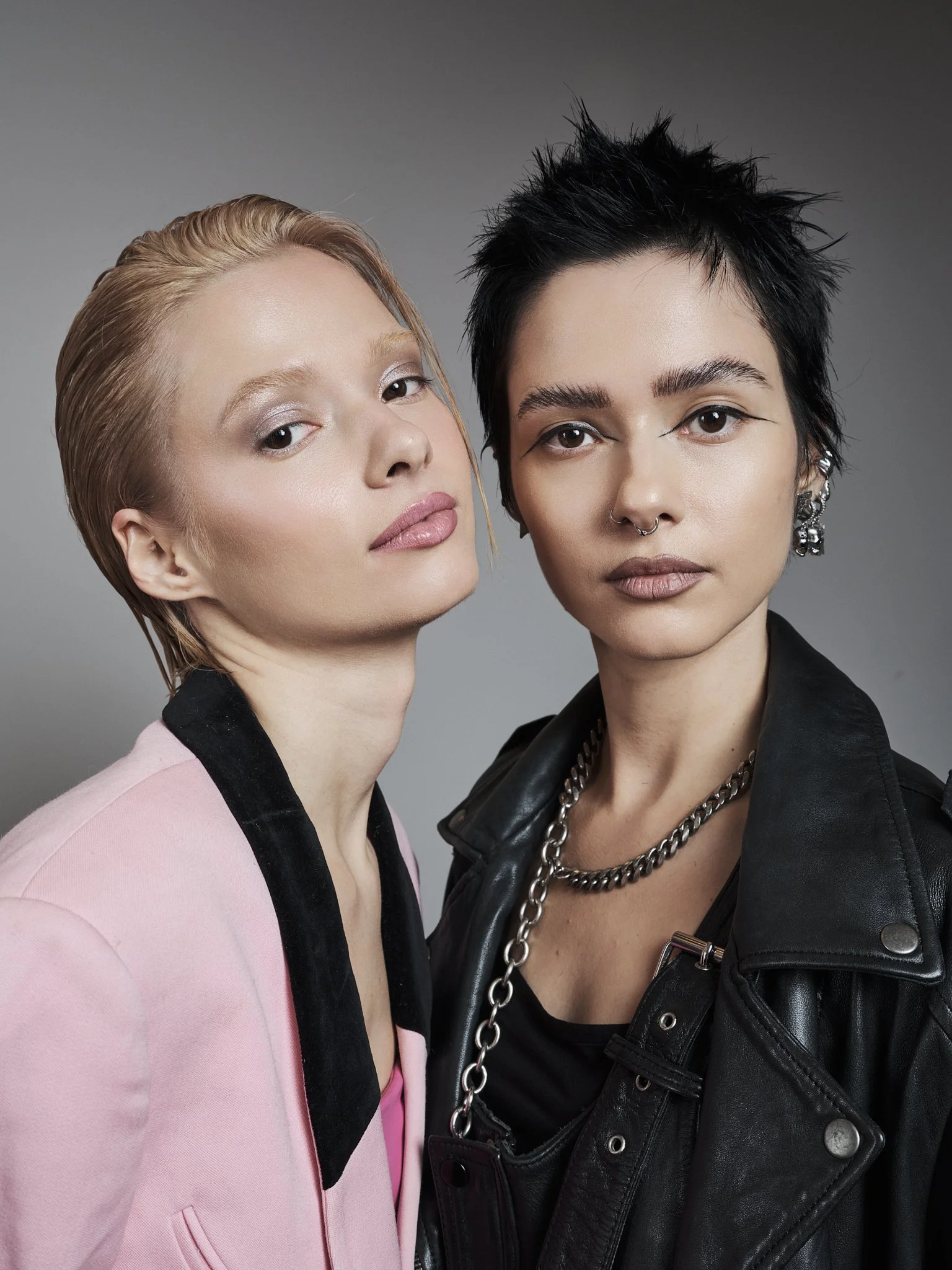 A portrait of the Bloom Twins, Two women standing close together, looking at the camera. One with short spiky black hair, wearing a black leather jacket and chain necklace, the other with short blonde hair, wearing a pink blazer with black collar.
